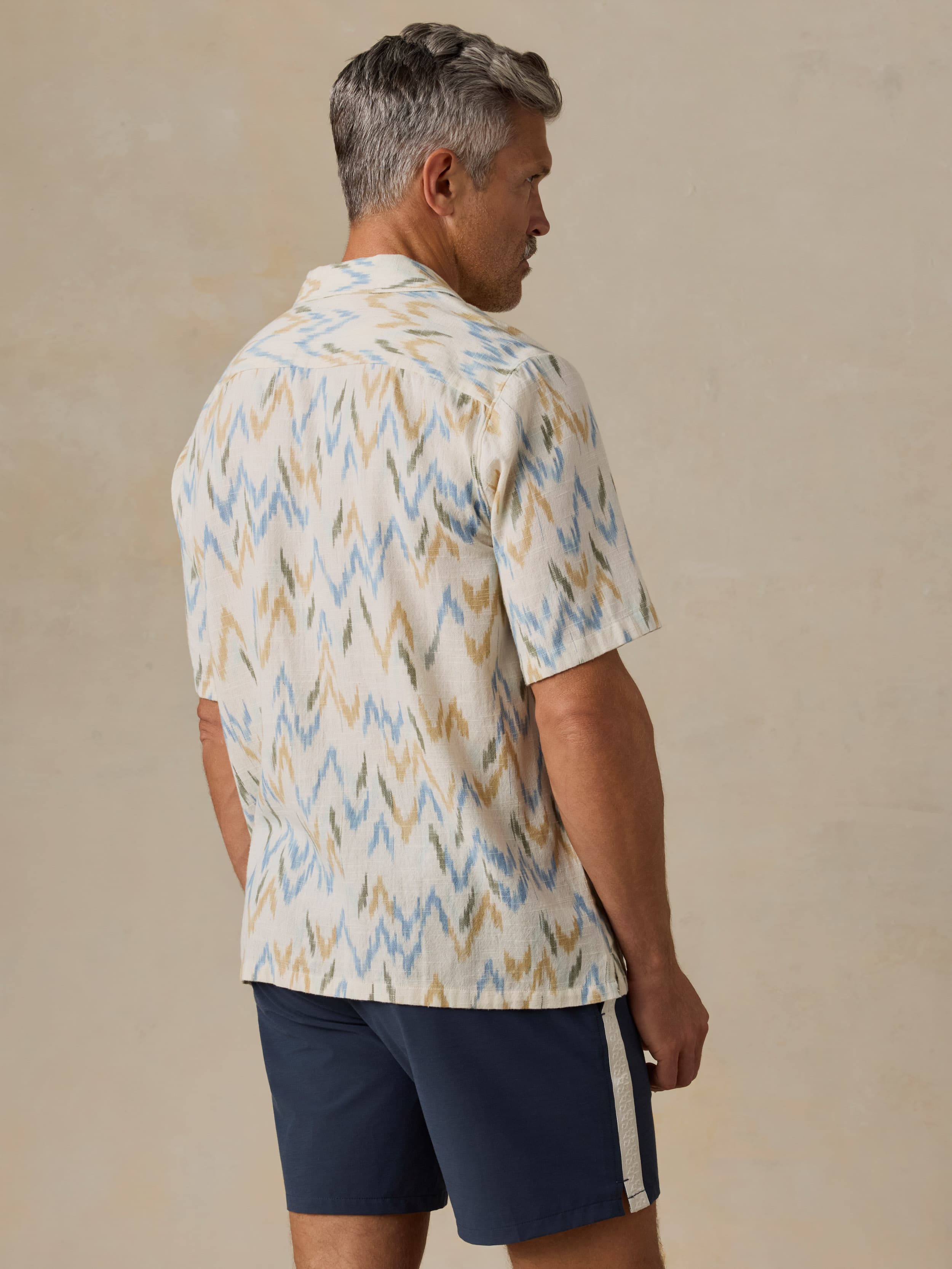 Man wearing a patterned shirt and navy shorts against a beige background