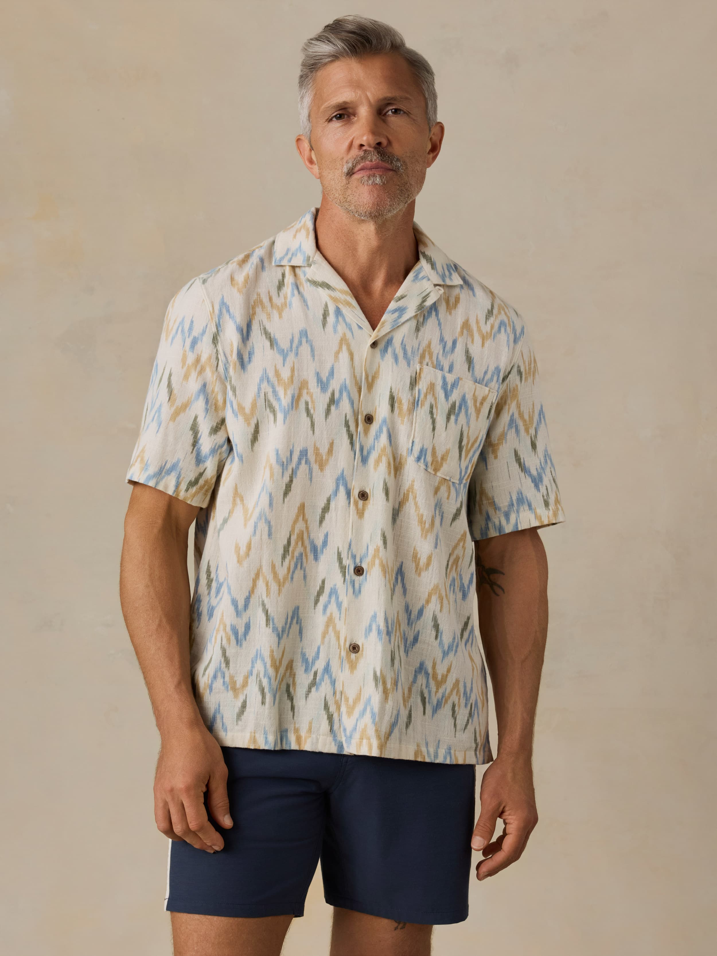 Man wearing a patterned shirt and navy shorts against a beige background