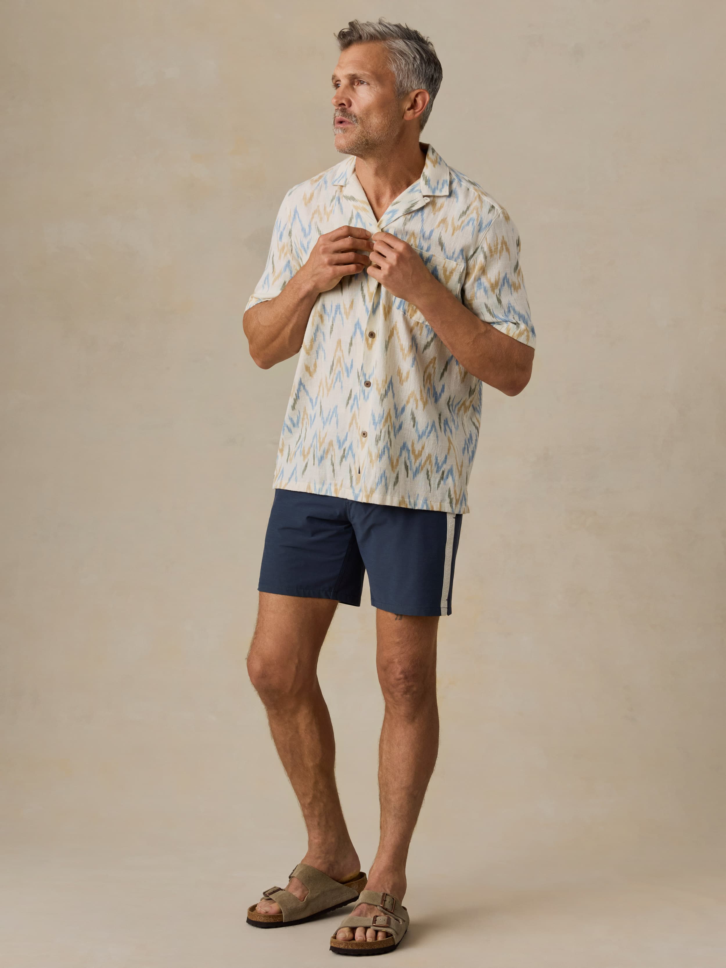 Man wearing a patterned shirt and navy shorts against a beige background