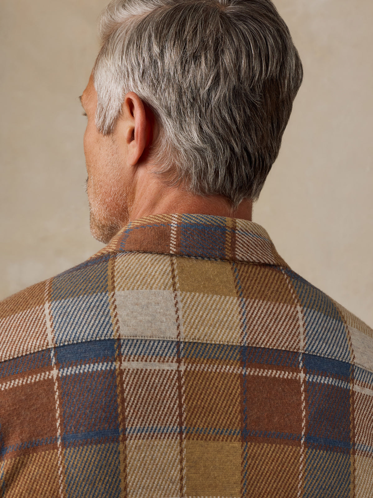 Close-up back view of a man wearing a brown, beige, and blue plaid wool shirt jacket against a neutral beige background., image 6 of 7