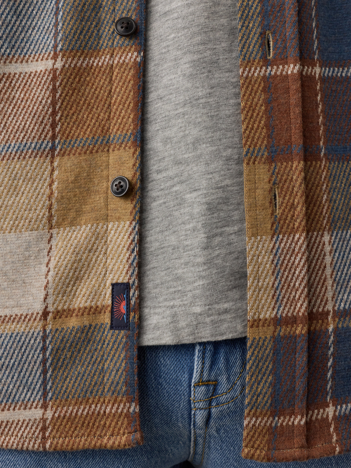 Close-up of a plaid overshirt in tan, brown, and blue tones layered over a heather gray T-shirt and blue jeans, with a small visible Faherty brand tab on the shirt., image 5 of 7