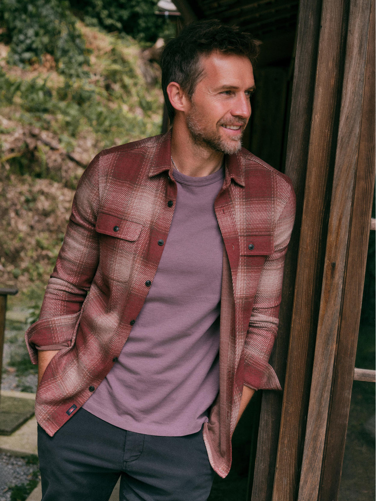 A man wearing a rust red and brown plaid flannel shirt over a mauve-purple t-shirt, standing outdoors next to a wooden structure., image 1 of 7