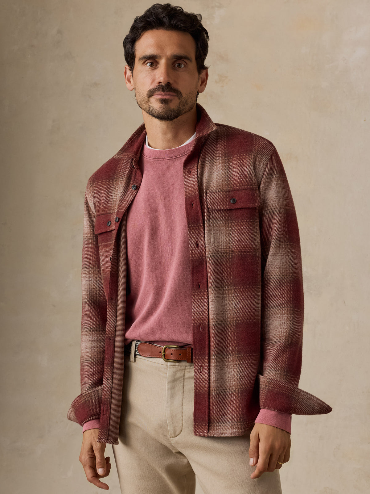 A man wearing a burgundy and brown plaid button-up overshirt over a dusty pink t-shirt, paired with beige trousers and a brown belt, standing against a textured beige wall., image 2 of 7