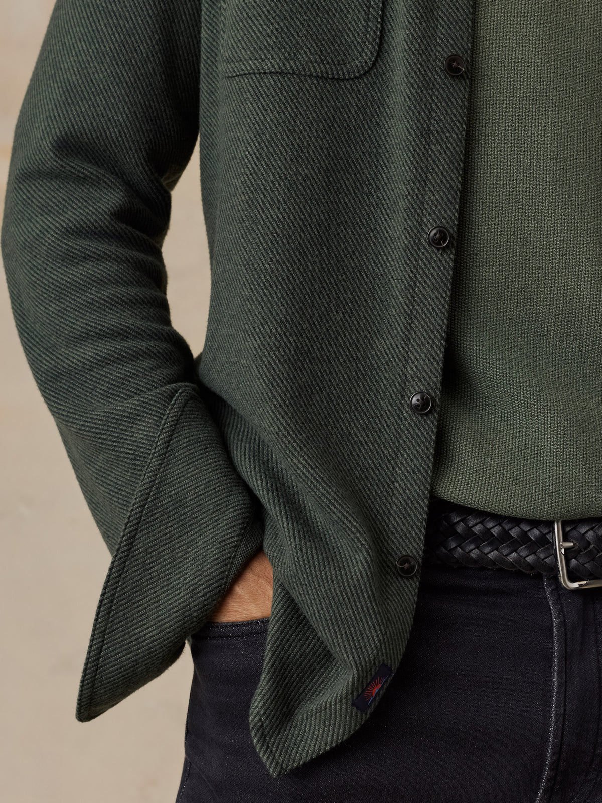 Close-up of a man wearing a dark green textured overshirt over an olive green knit sweater, paired with dark jeans and a black braided belt., image 3 of 5
