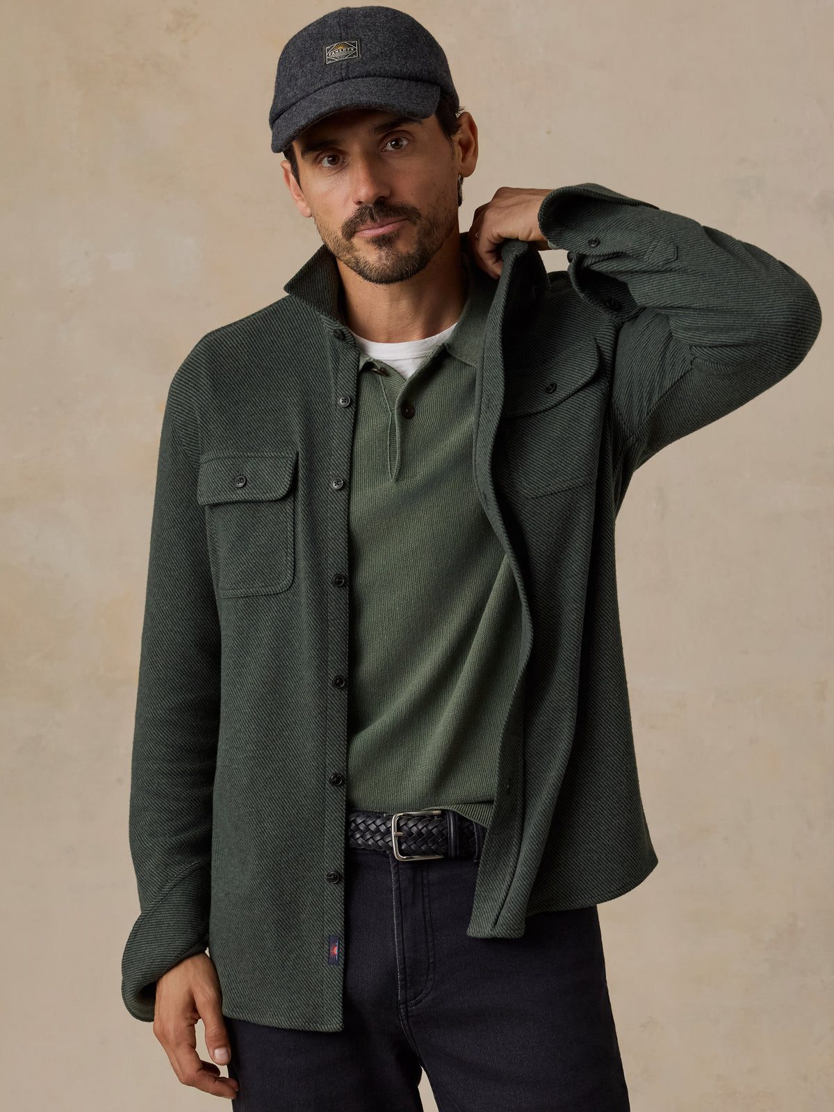 A man wearing a dark olive green button-up overshirt over a matching green henley shirt, dark jeans, and a dark gray baseball cap, posed against a textured beige background., image 1 of 5