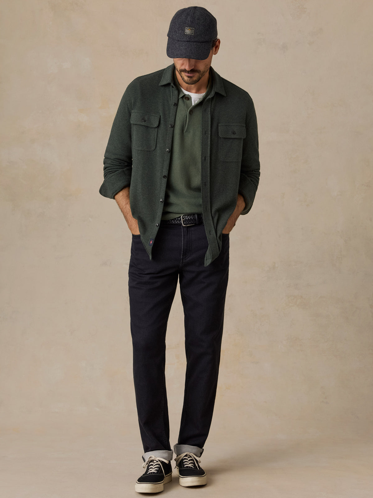A man wearing a dark green overshirt over an olive green t-shirt, dark navy pants, black sneakers, and a dark cap, standing against a textured beige studio backdrop., image 2 of 5