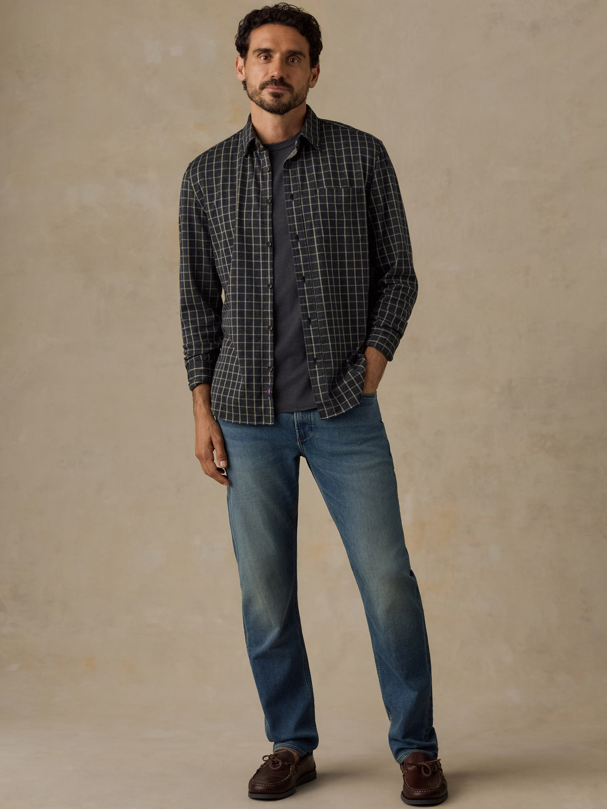 A man modeling a dark gray and black plaid button-down shirt over a dark t-shirt, paired with blue jeans and brown leather shoes, standing against a textured beige studio backdrop., image 6 of 7