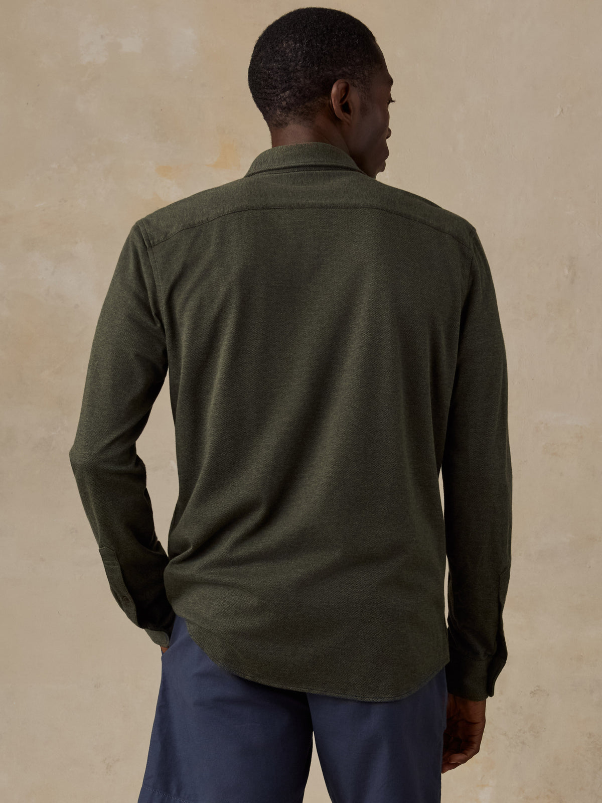 A man viewed from behind wearing an olive green long-sleeve button-up shirt and navy blue trousers, standing against a textured beige wall., image 3 of 5