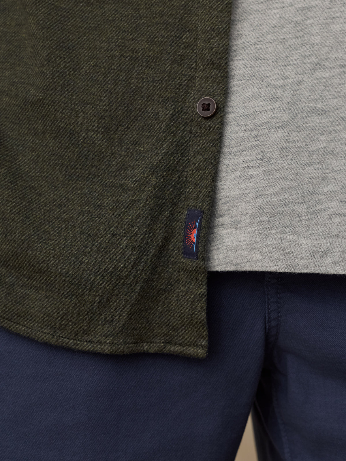 Close-up of a man wearing an olive green textured overshirt over a light gray heathered t-shirt and navy blue trousers, with a small Faherty brand tag visible on the overshirt., image 5 of 5
