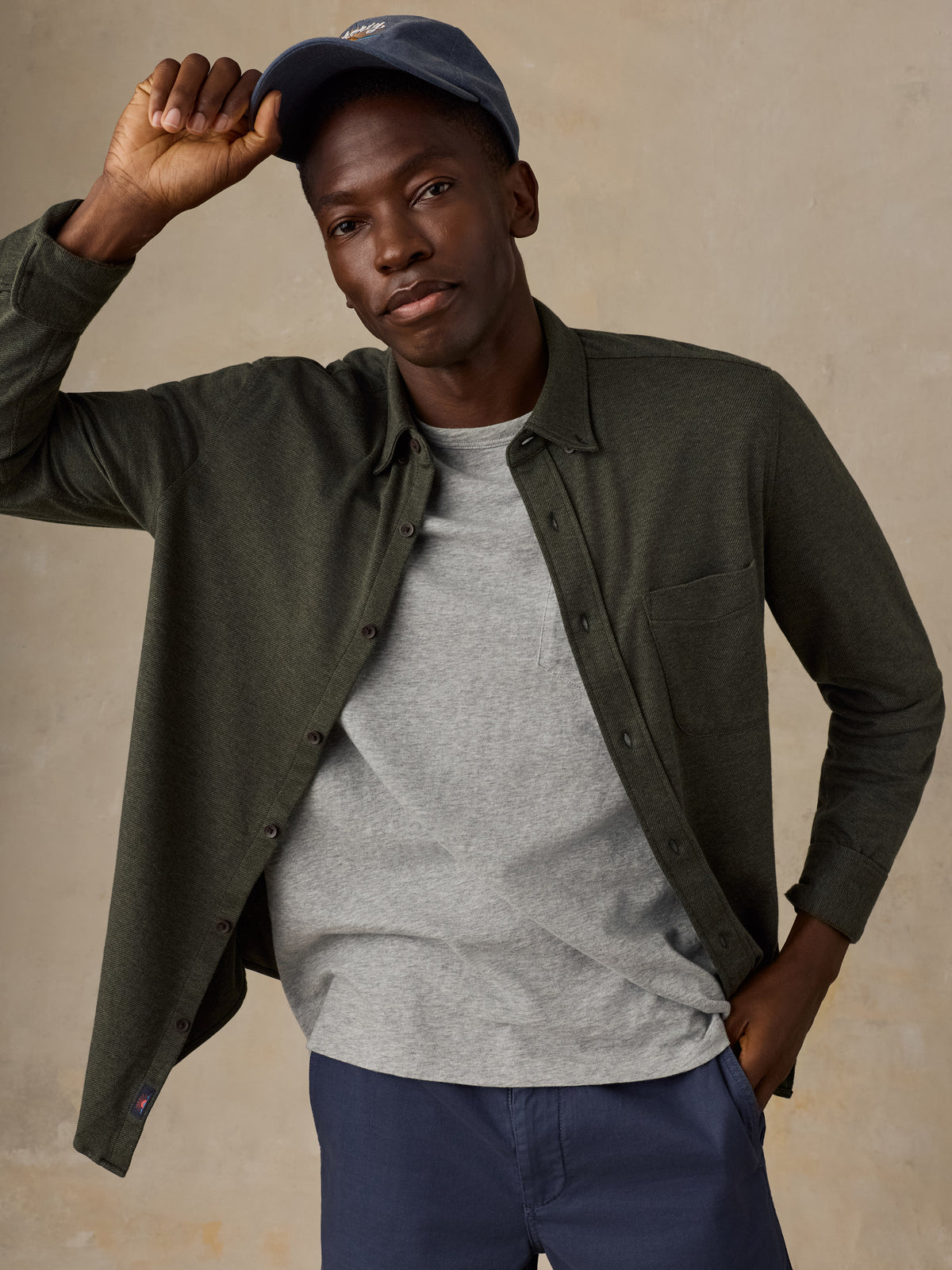 A man wearing an olive green long-sleeve button-up shirt over a heather gray t-shirt, paired with navy blue pants and a navy baseball cap, posing against a textured beige background., image 4 of 5