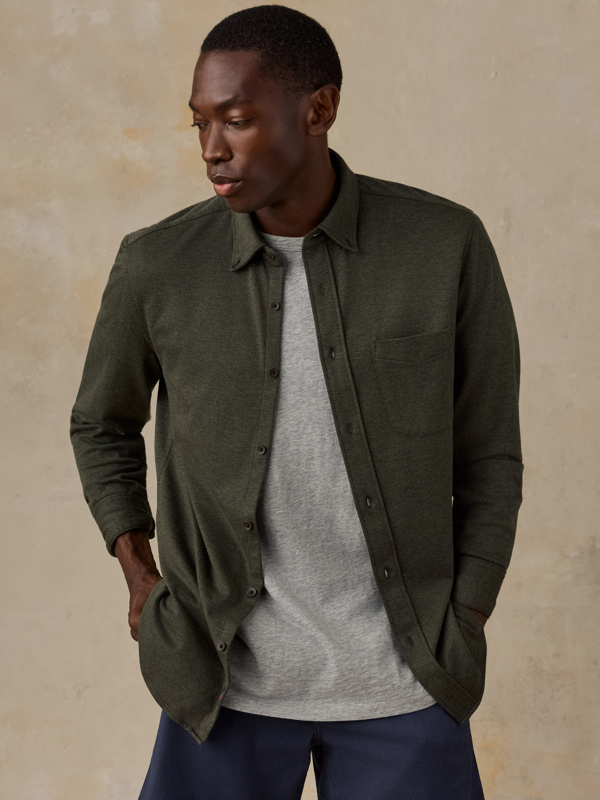 A man wearing an olive green long-sleeve button-up shirt over a light gray t-shirt and navy blue pants, standing against a textured beige background., image 1 of 5