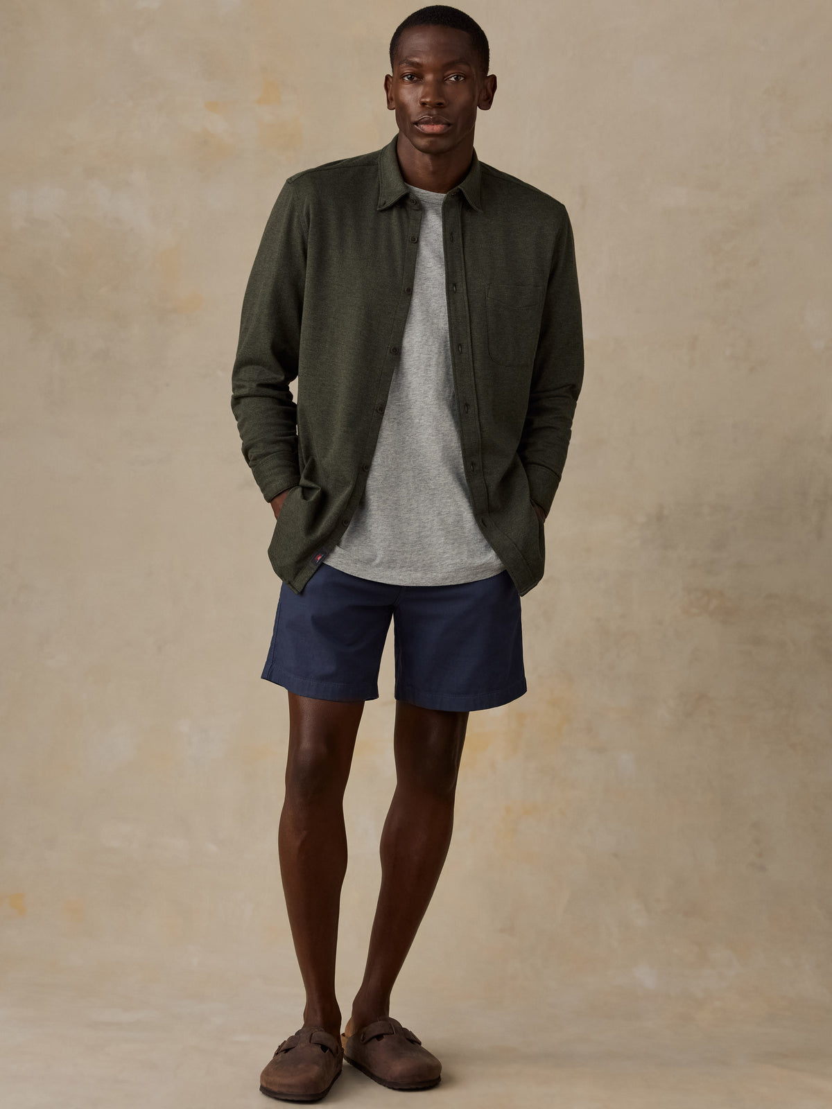 A man stands against a textured beige studio backdrop wearing an olive green button-up shirt over a light gray crewneck sweater, navy blue shorts, and brown slip-on shoes., image 2 of 5
