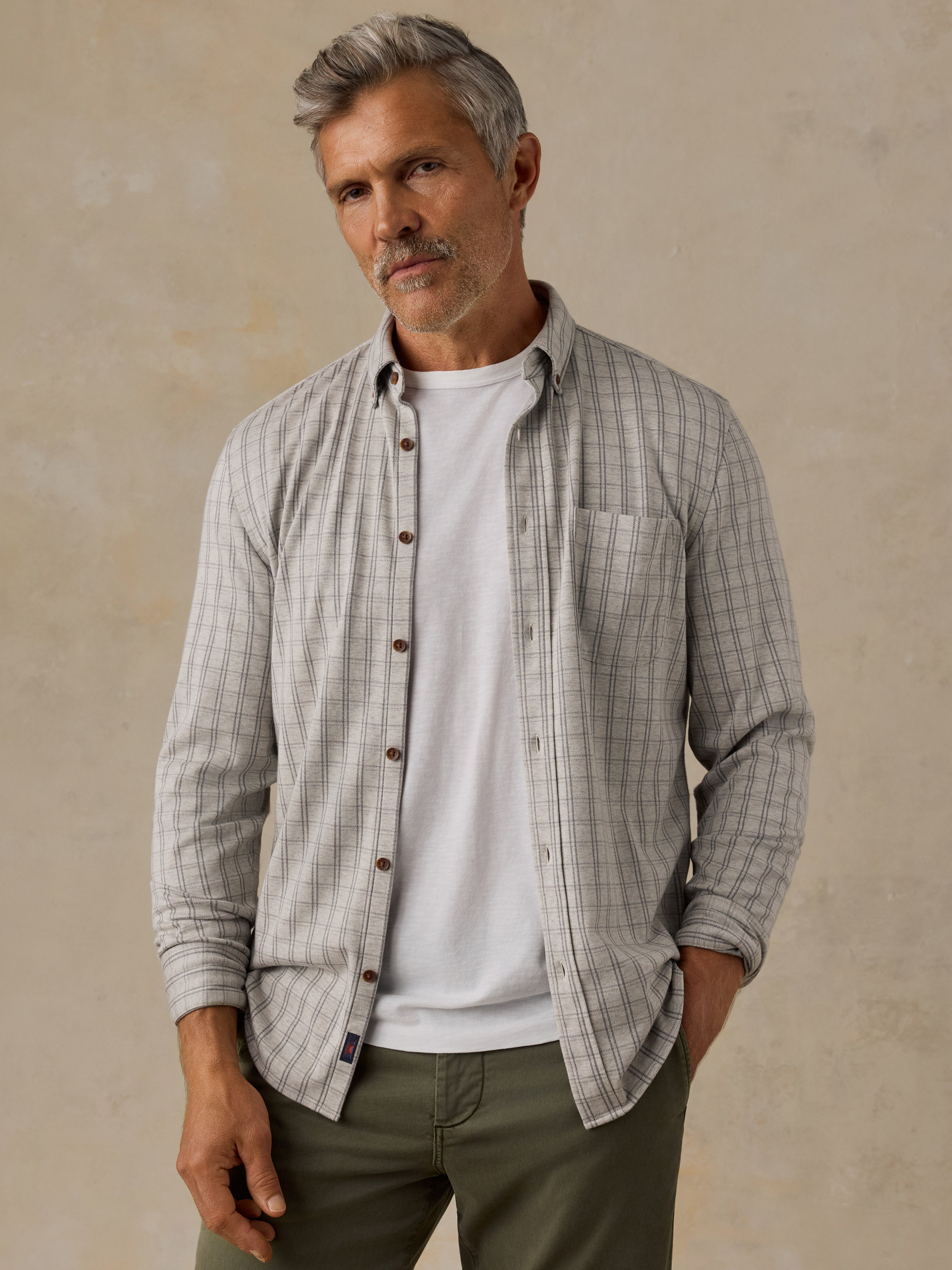 A man wearing a light gray and white plaid button-down shirt over a white t-shirt and olive green pants, standing against a textured beige background.
