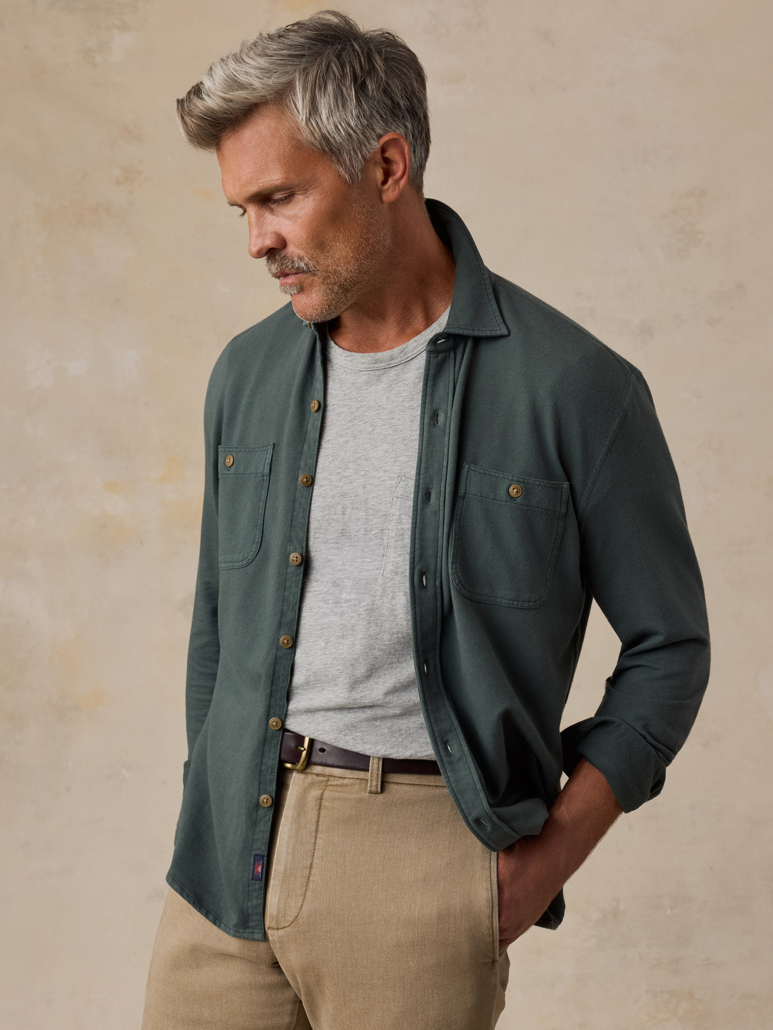 A man wearing a dark green button-up shirt over a light gray t-shirt, paired with beige trousers, standing against a textured beige background.