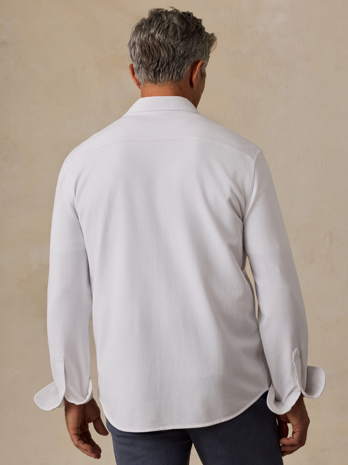 A man viewed from behind wearing a white long-sleeve button-down shirt and dark trousers, standing against a textured beige wall., image 4 of 6