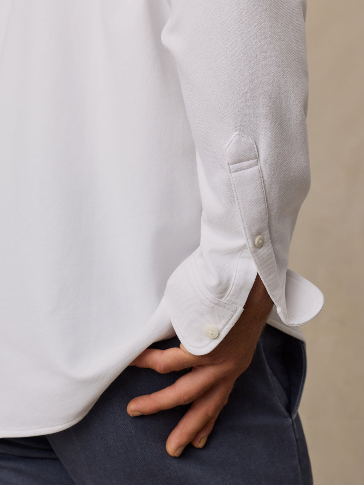 Close-up of a person wearing a white long-sleeve button-up shirt with the cuff turned up, paired with dark navy trousers, against a neutral beige background., image 5 of 6