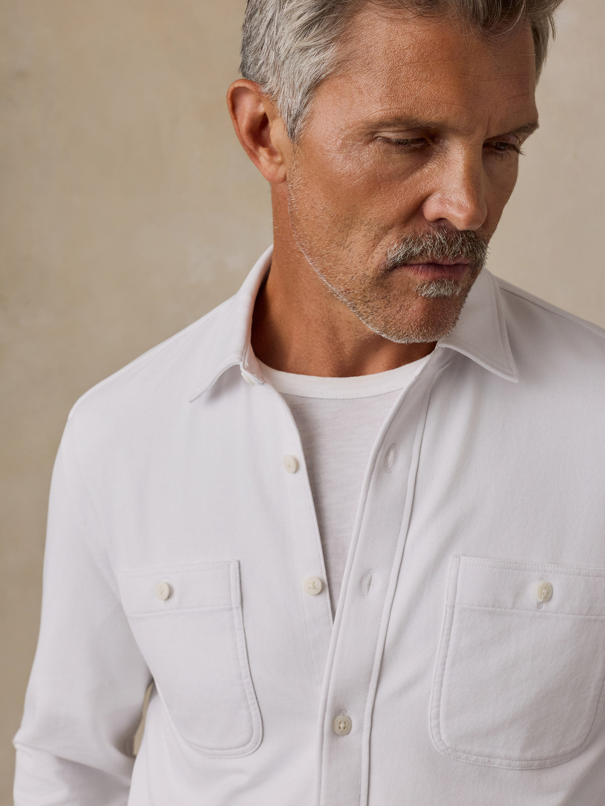 A close-up of a man wearing a white long-sleeve button-up shirt over a white t-shirt, set against a neutral beige background., image 2 of 6