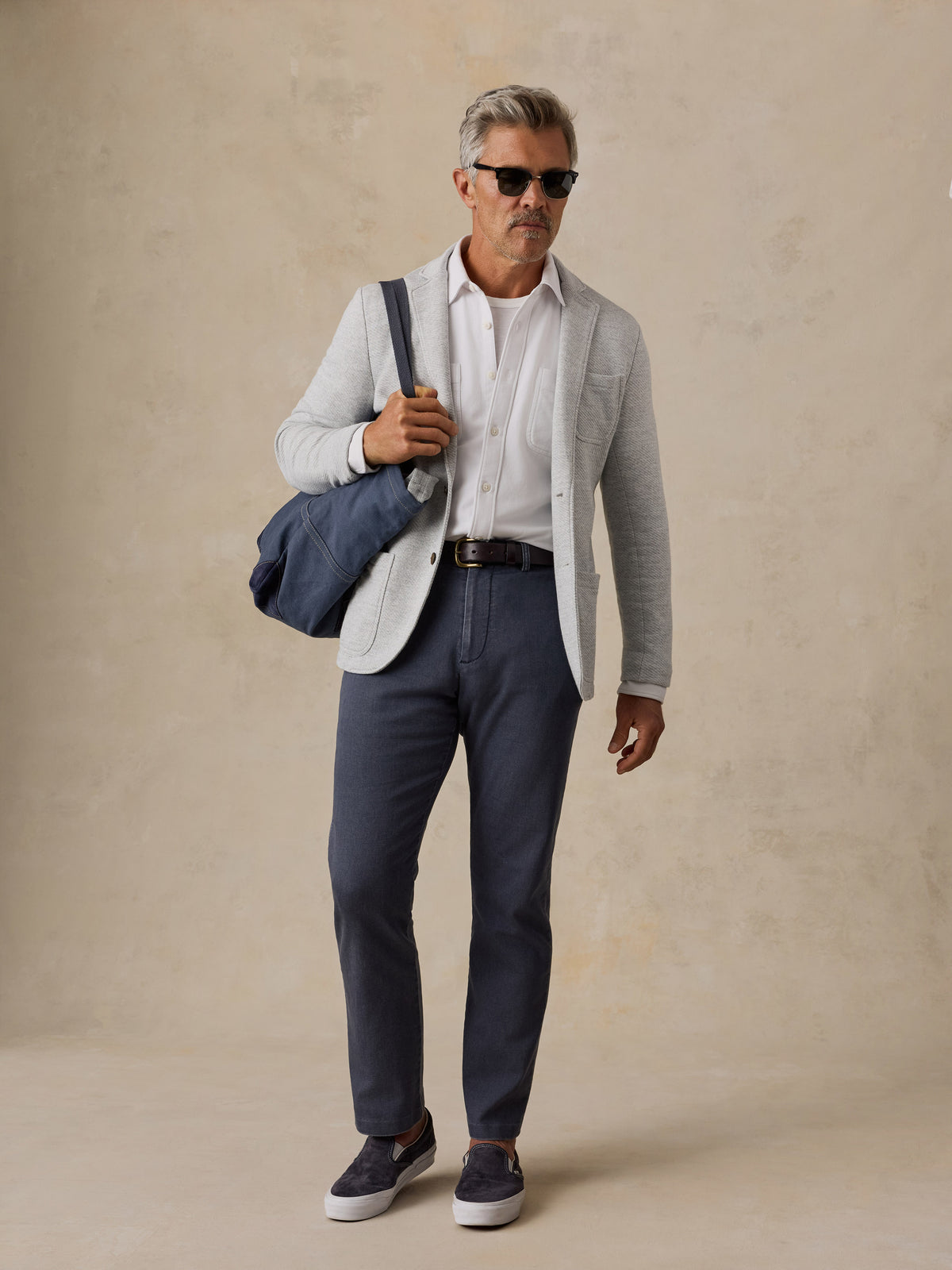 A man wearing a light gray blazer, white dress shirt, navy trousers, and navy slip-on shoes, carrying a navy duffel bag over his shoulder against a textured beige studio backdrop., image 6 of 6