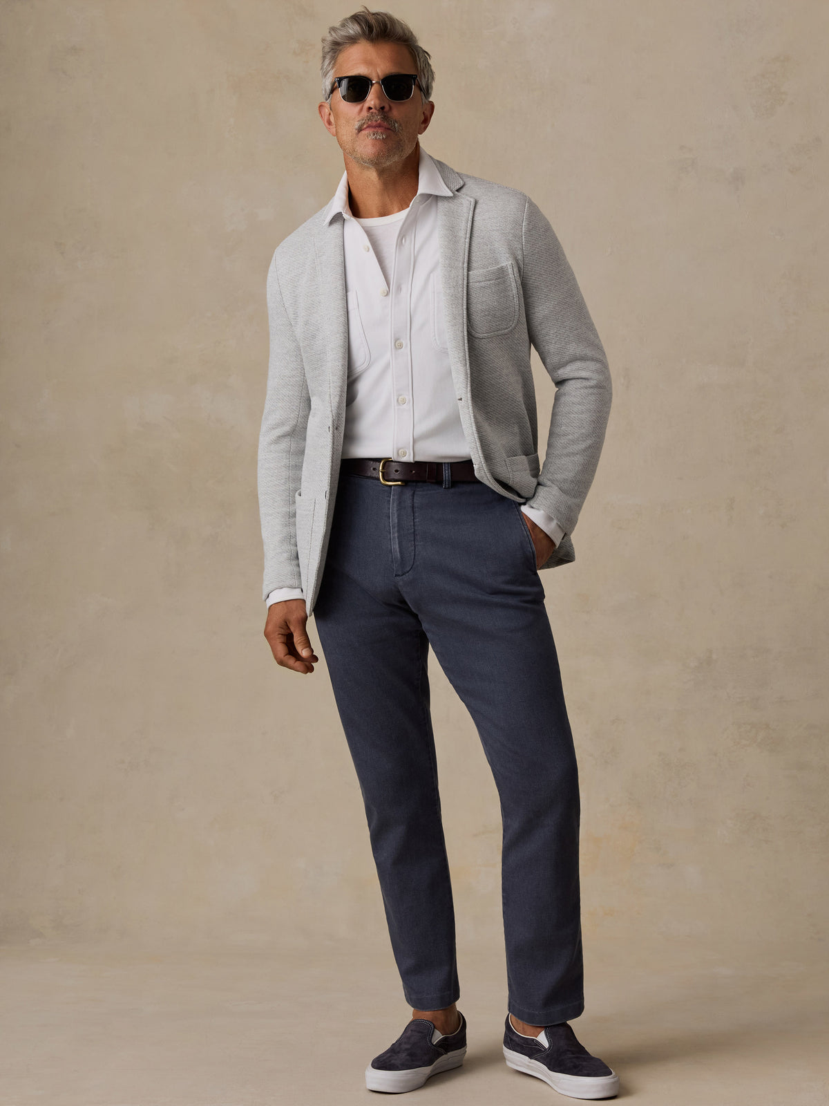 A man wearing a light gray knit cardigan over a white button-down shirt, navy blue trousers, and dark slip-on sneakers, standing against a textured beige studio backdrop., image 3 of 6