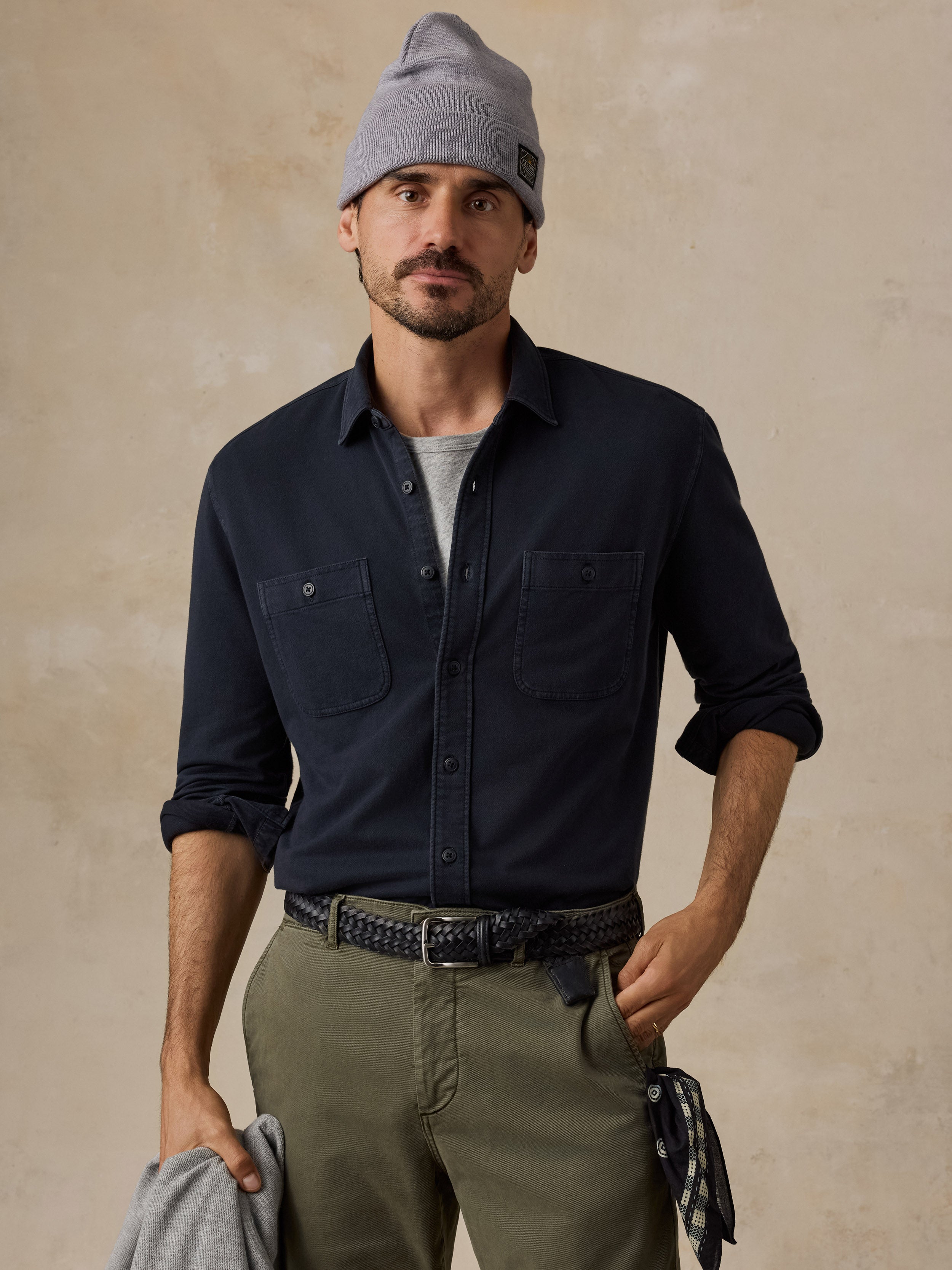 A man wearing a dark navy long-sleeve button-up shirt with chest pockets, olive green pants, a gray beanie, and a black belt, standing against a textured beige background.