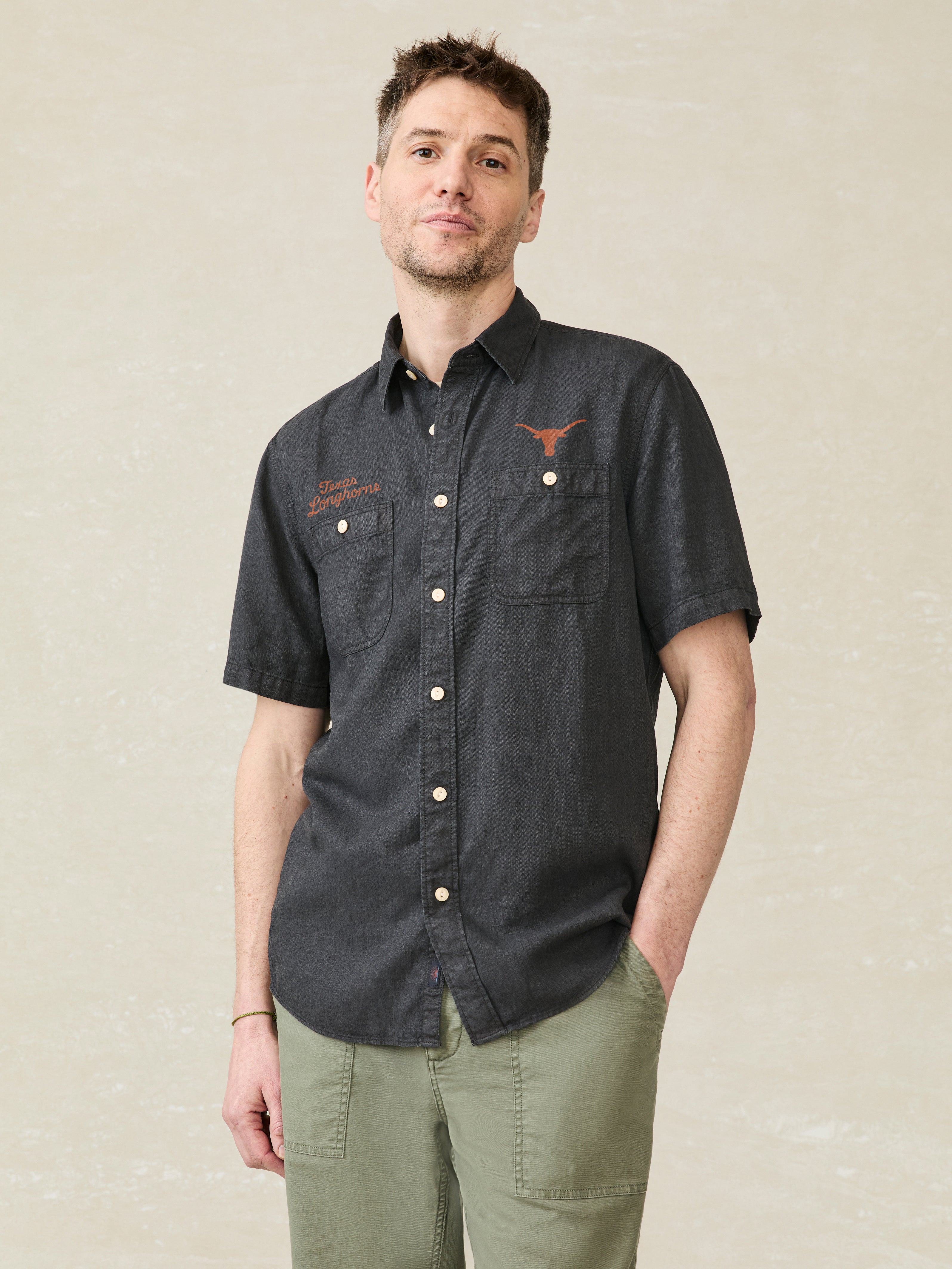 Man wearing a black short-sleeve shirt with an University of Texas logo above the left and right chest pockets against a beige background