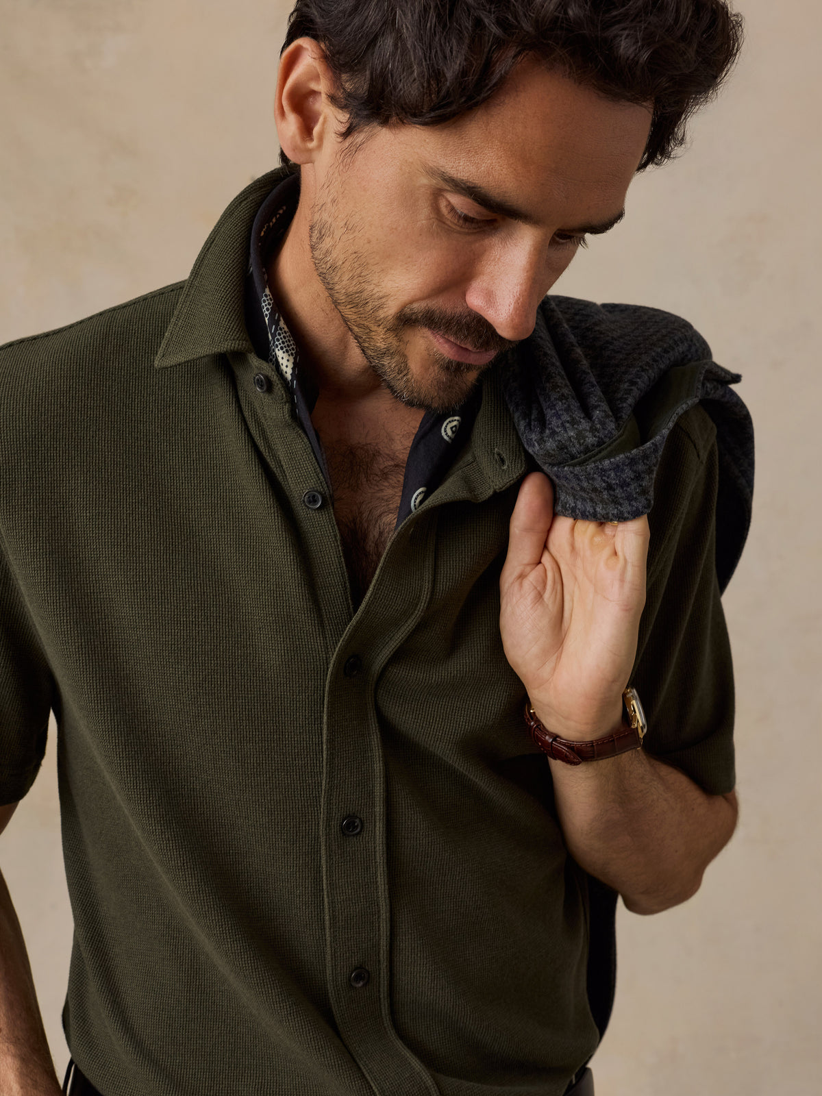 A man wearing an olive green short-sleeve button-up shirt, with a dark charcoal knit sweater draped over his shoulder, set against a warm beige textured background., image 2 of 6