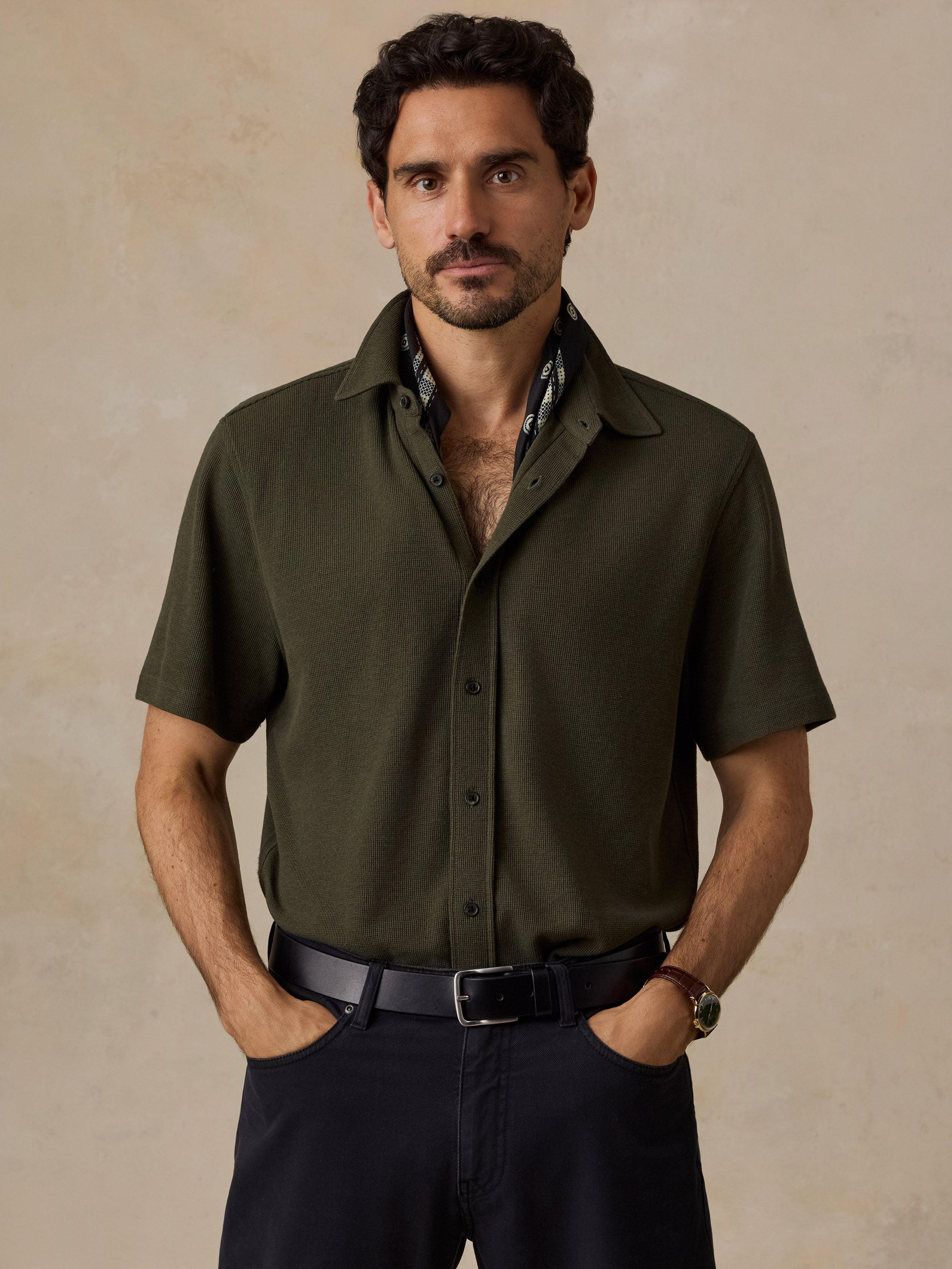 A man wearing a short-sleeve button-up shirt in olive green, paired with dark trousers and a black belt, standing against a textured beige background.