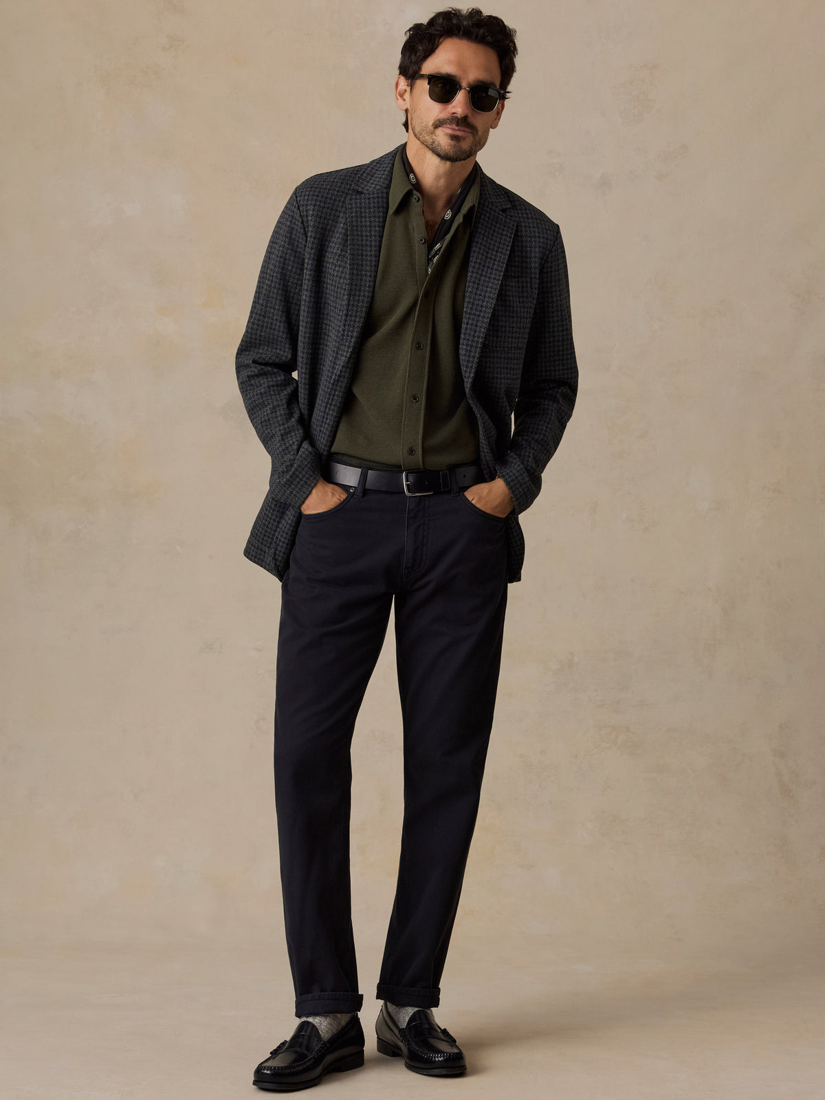 A man wearing a dark gray patterned blazer, olive green shirt, navy trousers, black loafers, and sunglasses, standing against a textured beige studio backdrop., image 6 of 6