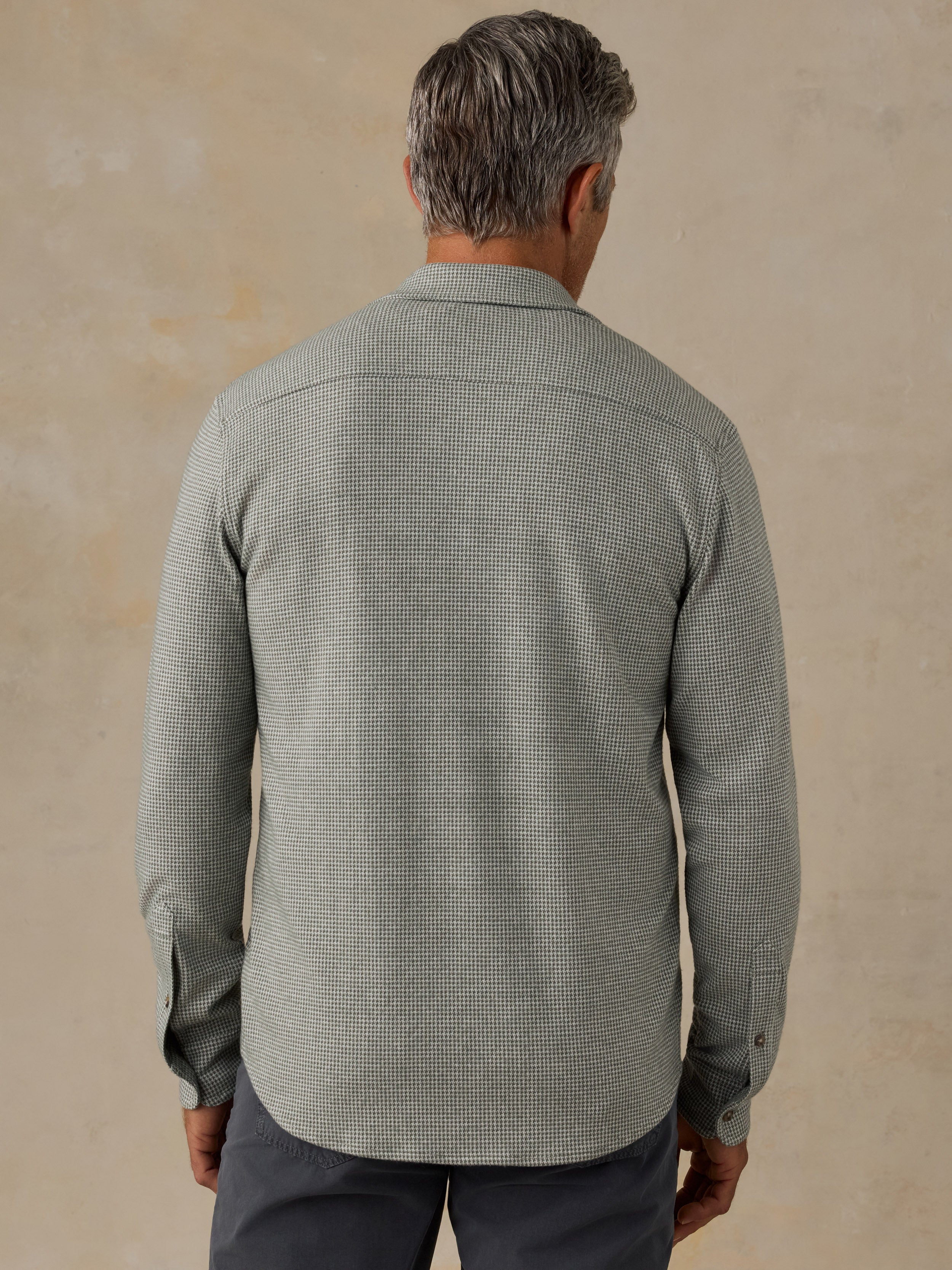 A man viewed from behind wearing a light sage green and white micro-check patterned long-sleeve shirt, paired with dark gray trousers, against a textured beige studio background.