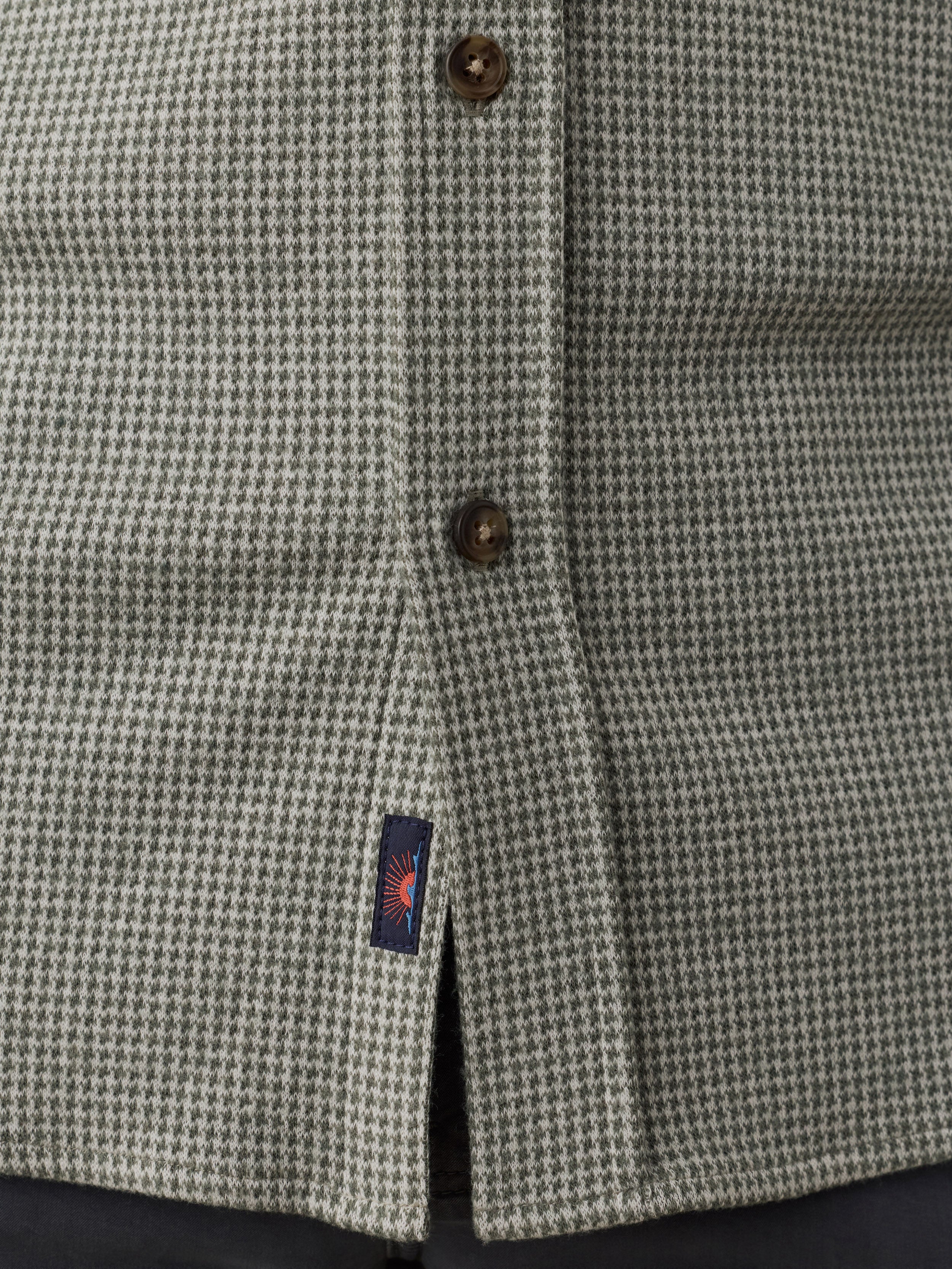 Close-up of a textured houndstooth patterned shirt in gray and white, featuring dark brown buttons and a small navy blue Faherty brand tag near the hem.