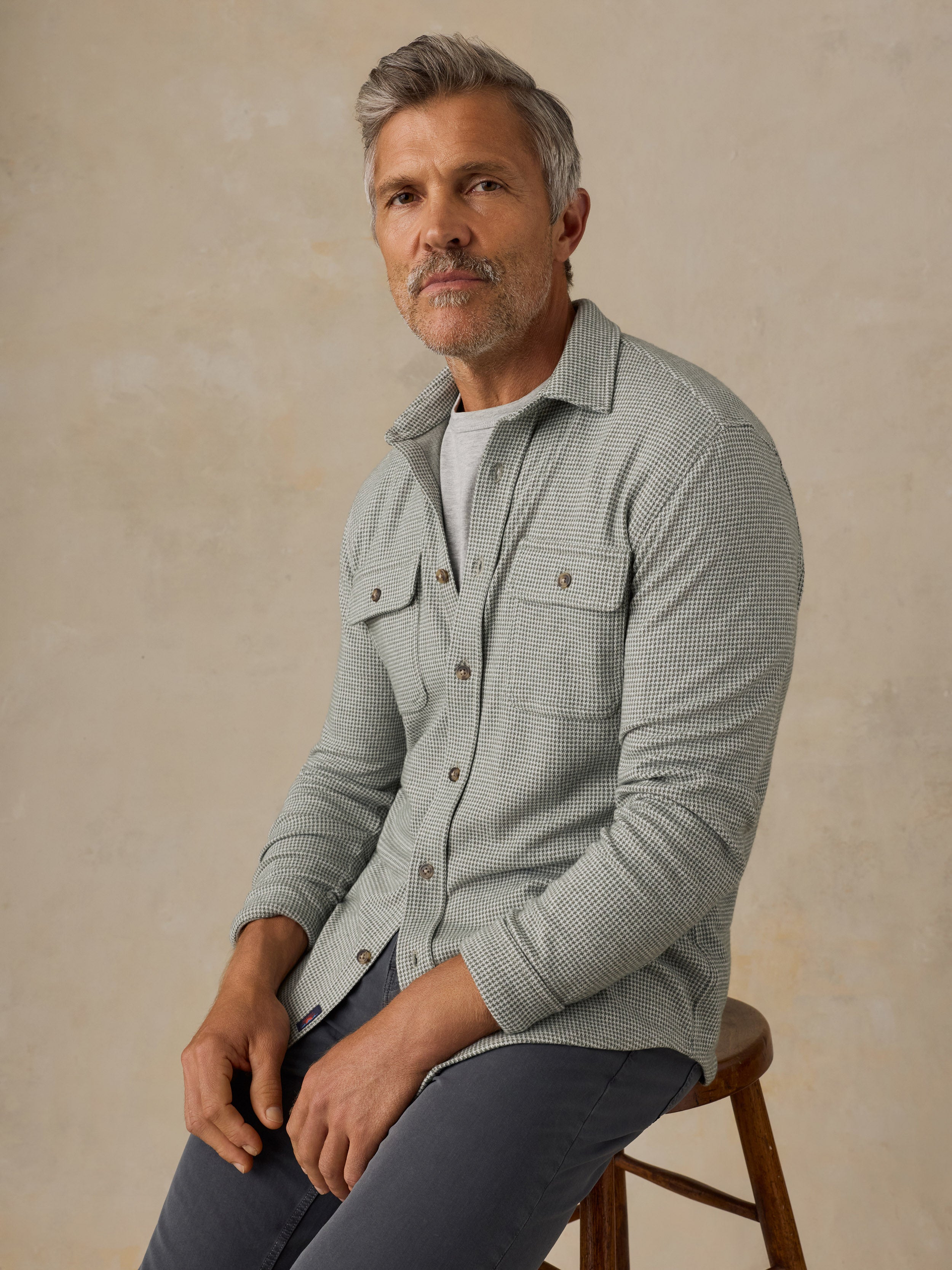 A man with gray hair and a beard sits on a wooden stool, wearing a light sage green textured button-down shirt over a white t-shirt and dark gray pants, against a textured beige background.