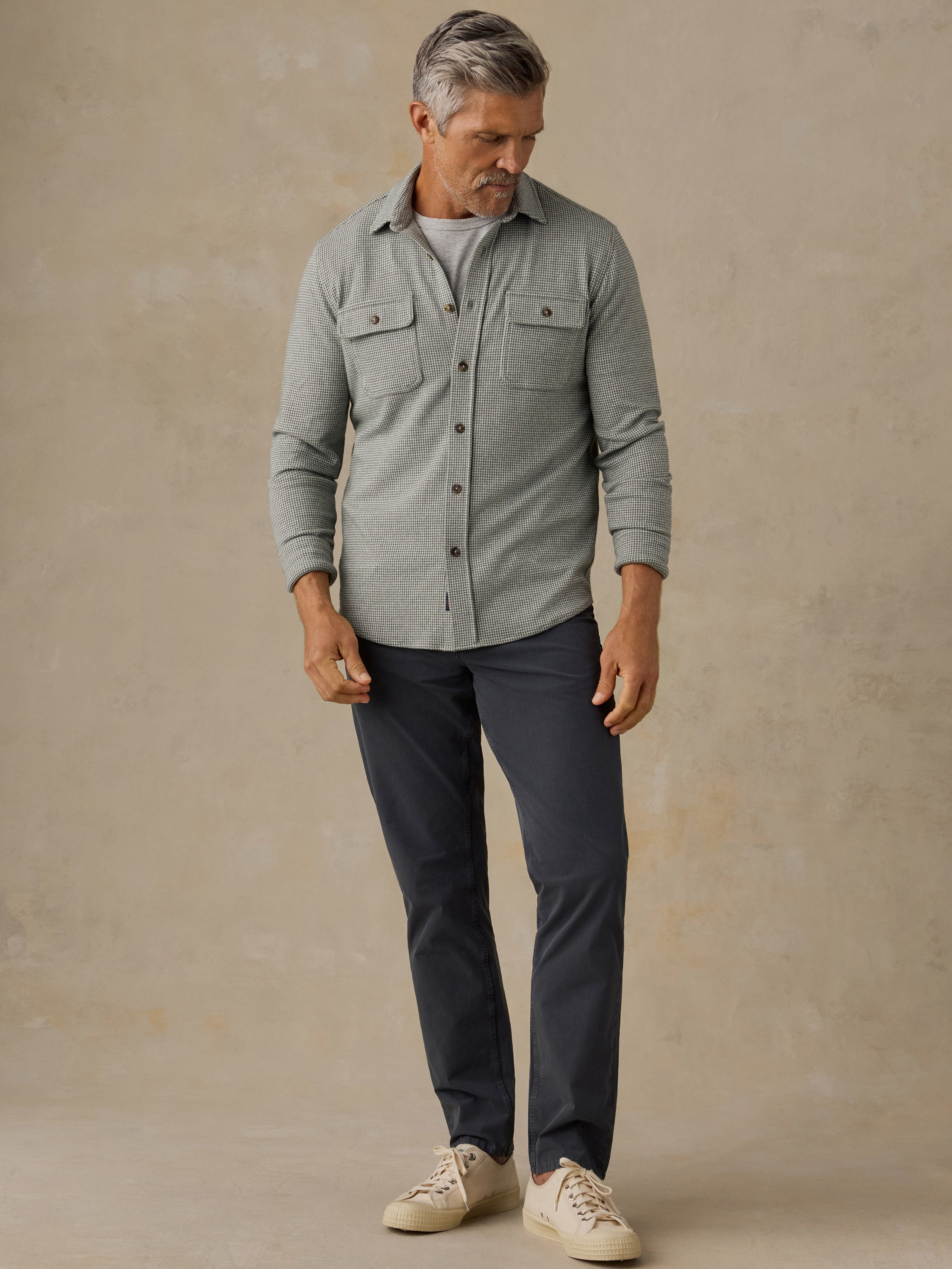A man wearing a light green long-sleeve button-up shirt, dark navy pants, and beige sneakers, standing against a textured beige studio backdrop.