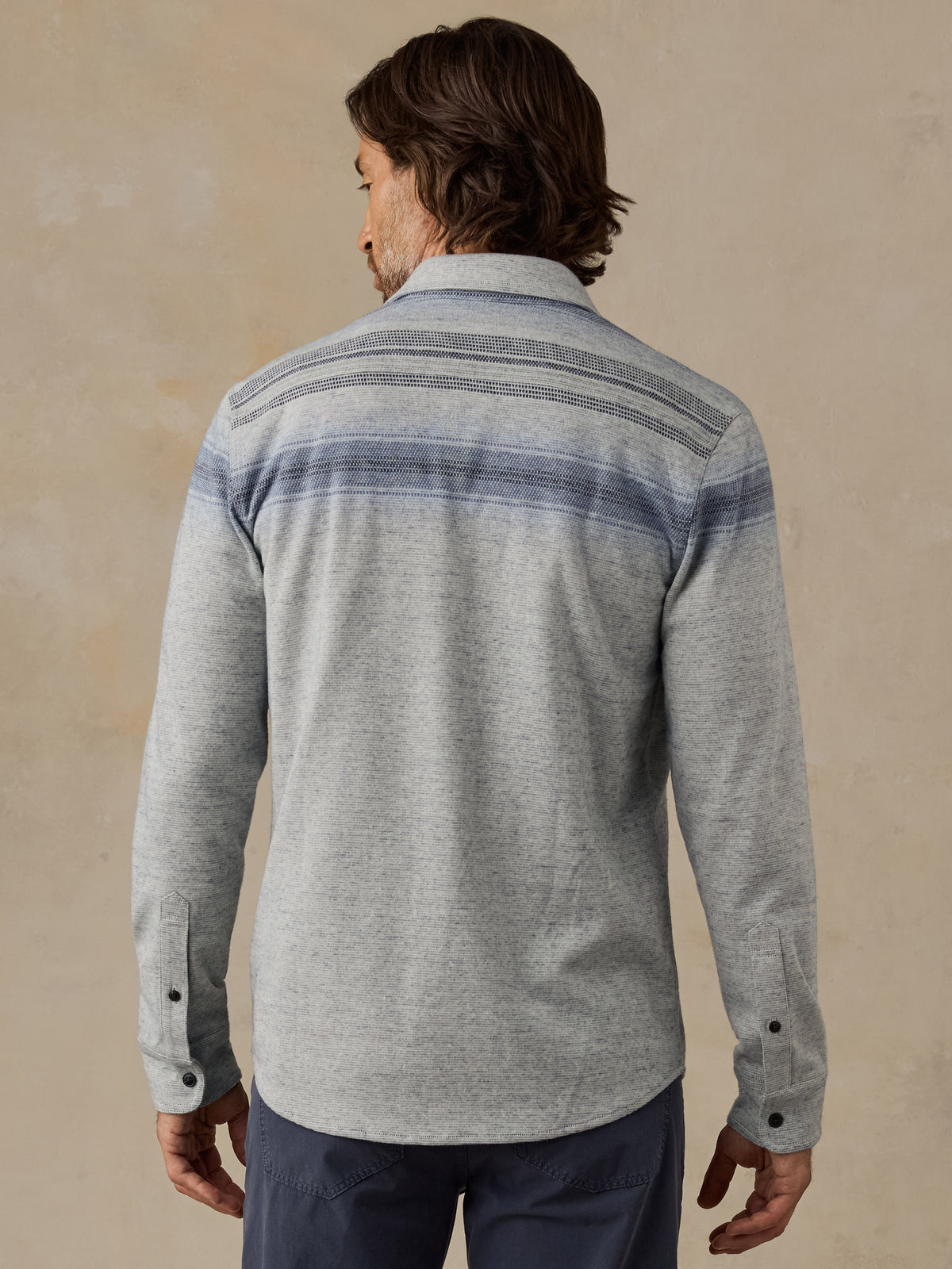 A man viewed from the back wearing a light gray long-sleeve button-up shirt with horizontal blue and gray stripes across the shoulders and upper back, paired with dark pants, against a textured beige background., image 3 of 7