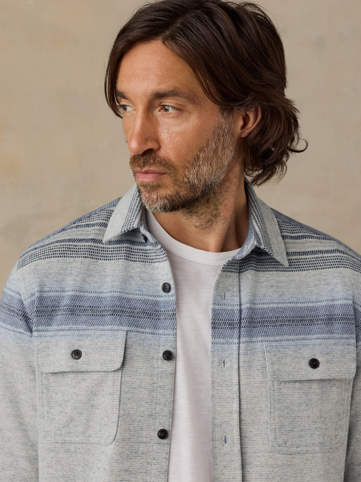 A man wearing a light gray and blue striped button-down shirt over a white t-shirt, photographed against a neutral beige background., image 5 of 7