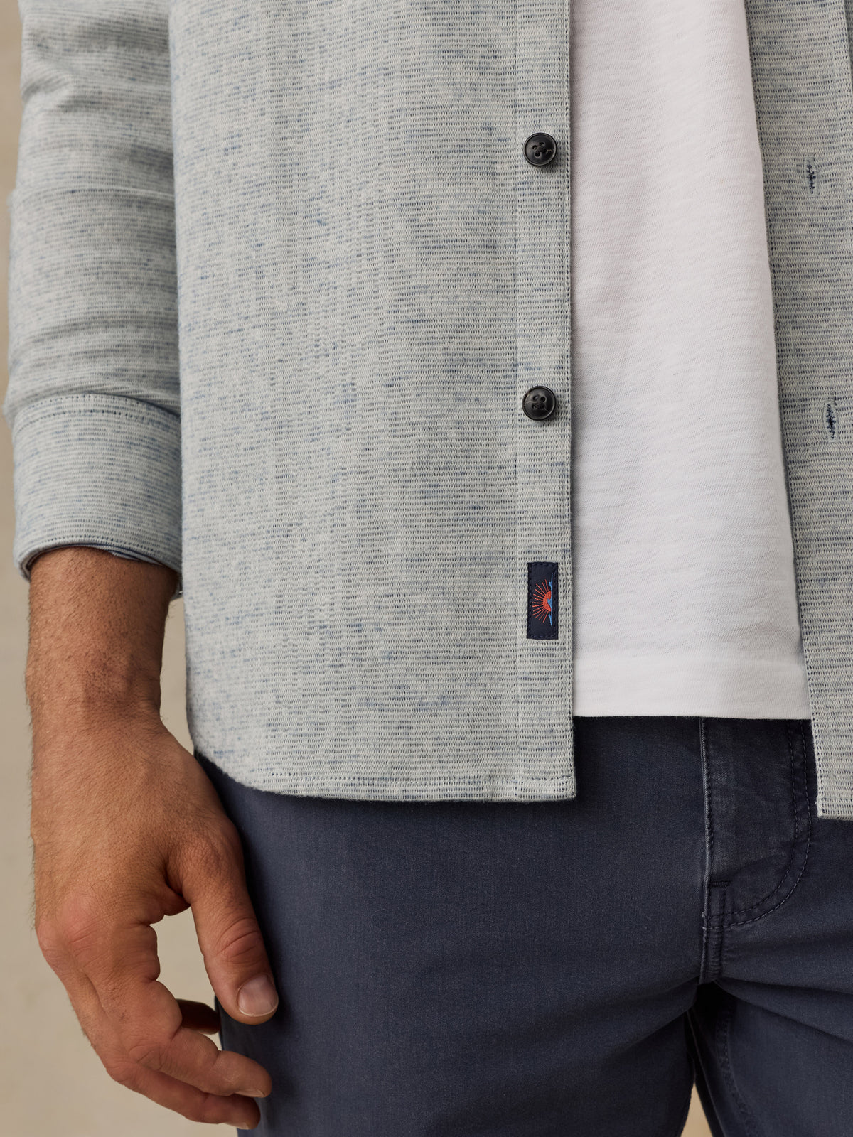 Close-up of a man wearing a light gray heathered knit cardigan over a white T-shirt and navy pants, with a small Faherty brand tag visible near the hem., image 4 of 7