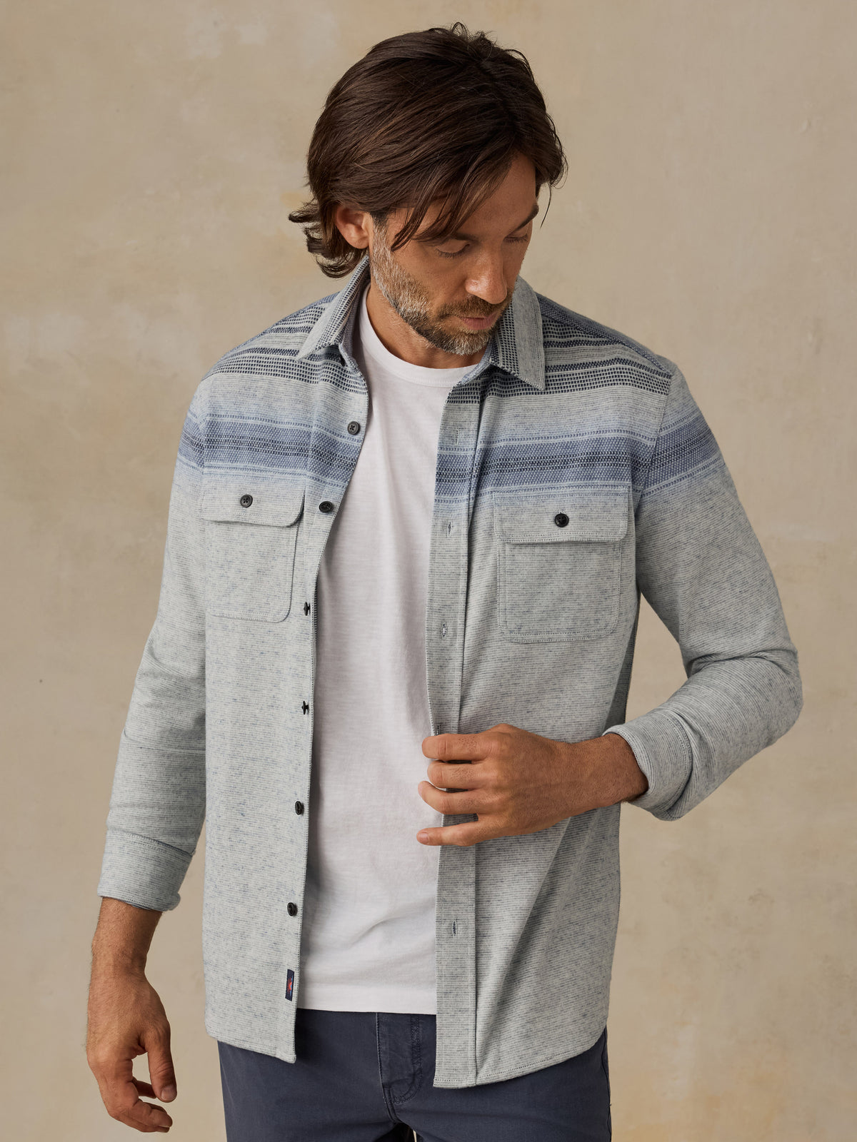 A man wearing a light gray heathered button-up shirt with blue and gray horizontal stripes across the chest, layered over a white t-shirt and dark blue jeans, standing against a textured beige background., image 1 of 7