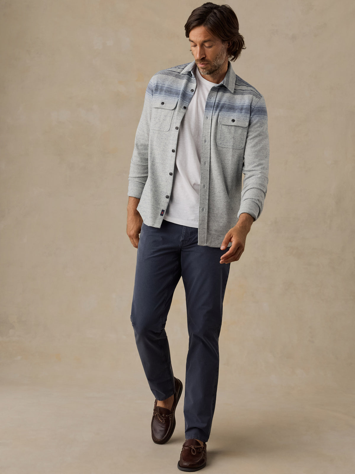 A man wearing a light gray and blue striped button-down shirt over a white t-shirt, navy blue pants, and brown leather loafers, standing against a textured beige studio backdrop., image 6 of 7