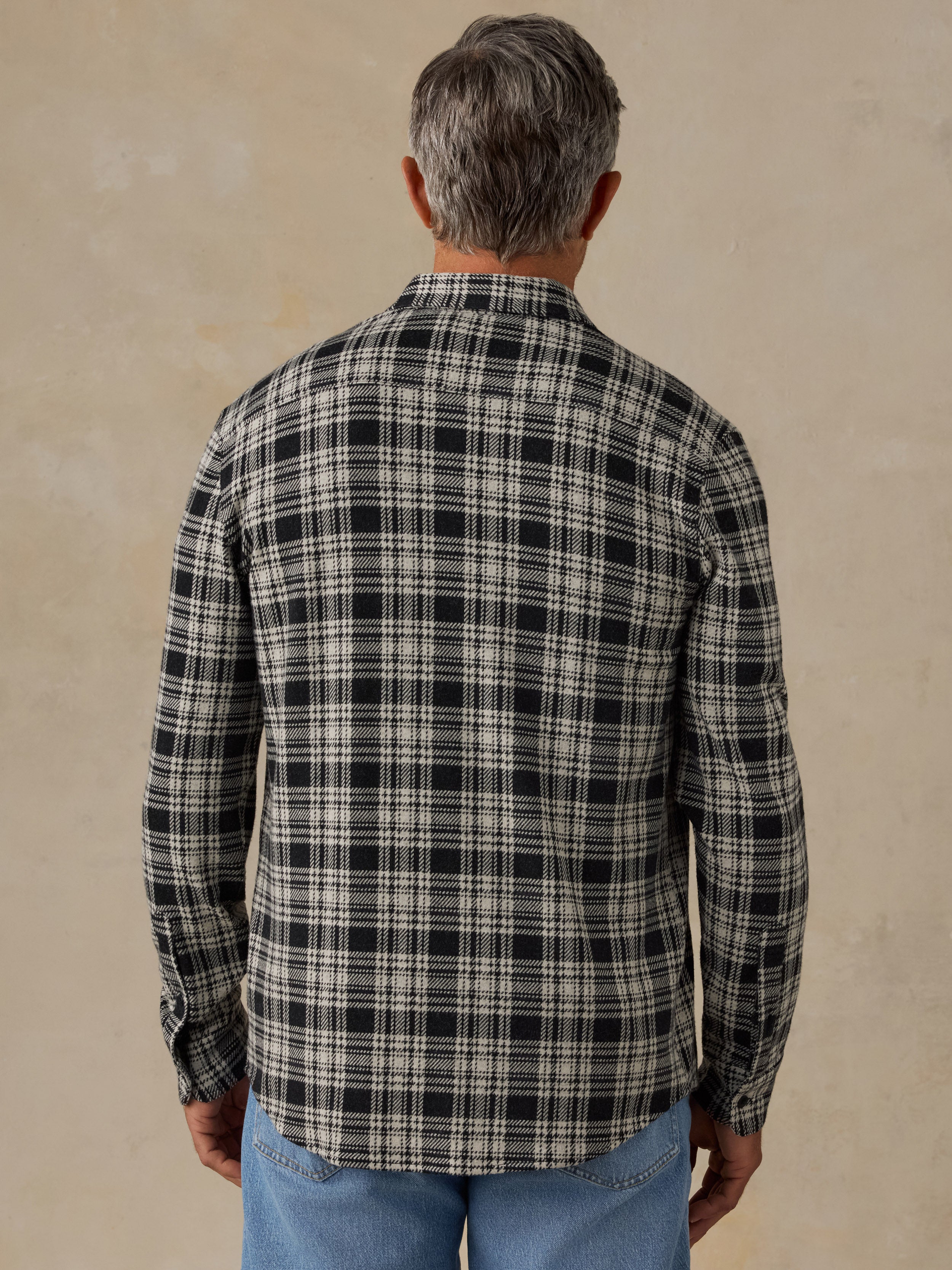 A man viewed from behind wearing a long-sleeve plaid button-up shirt in black and off-white, paired with blue jeans, against a textured beige background.