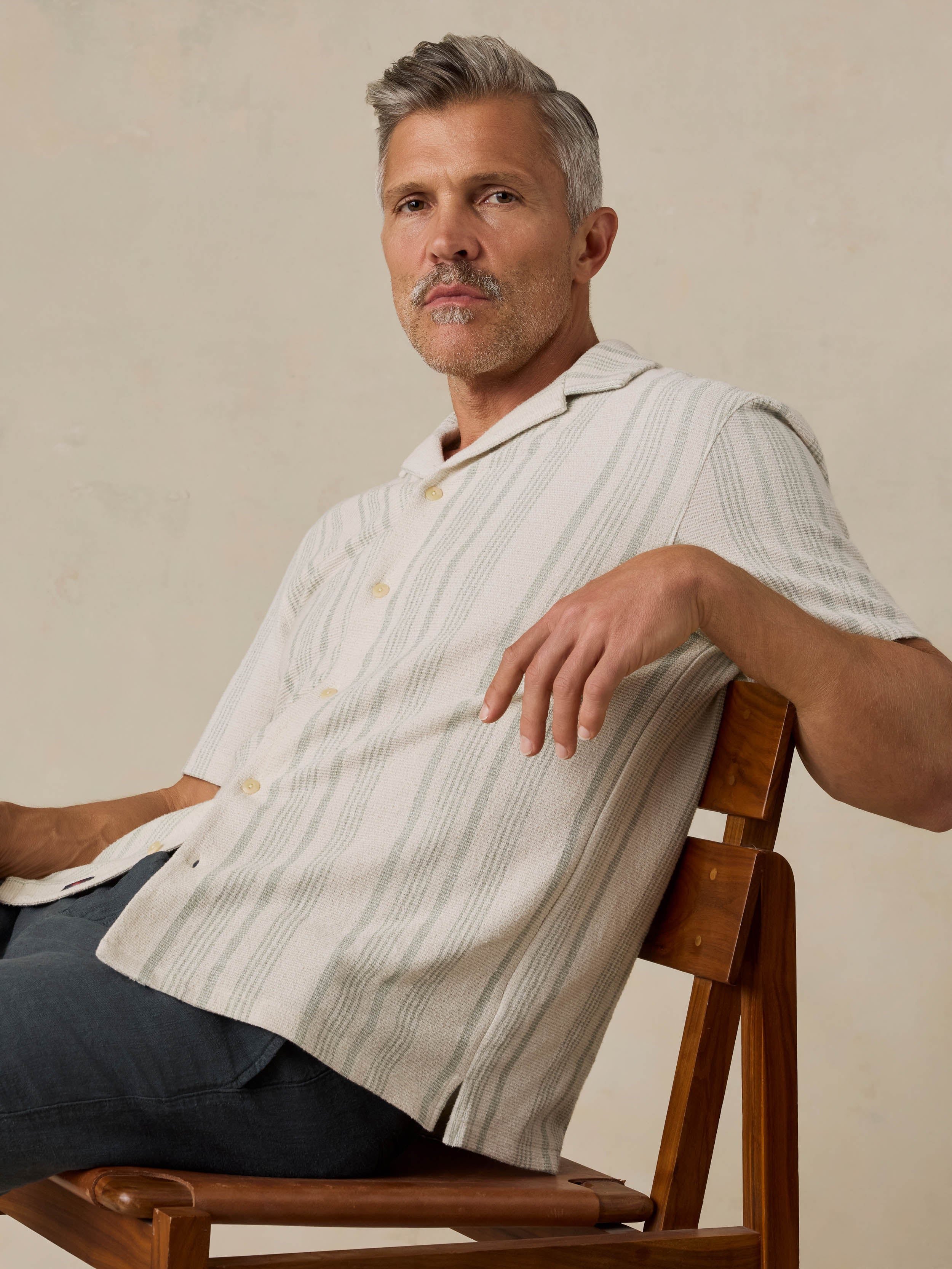 Man sitting on a wooden chair wearing a beige striped shirt against a beige background