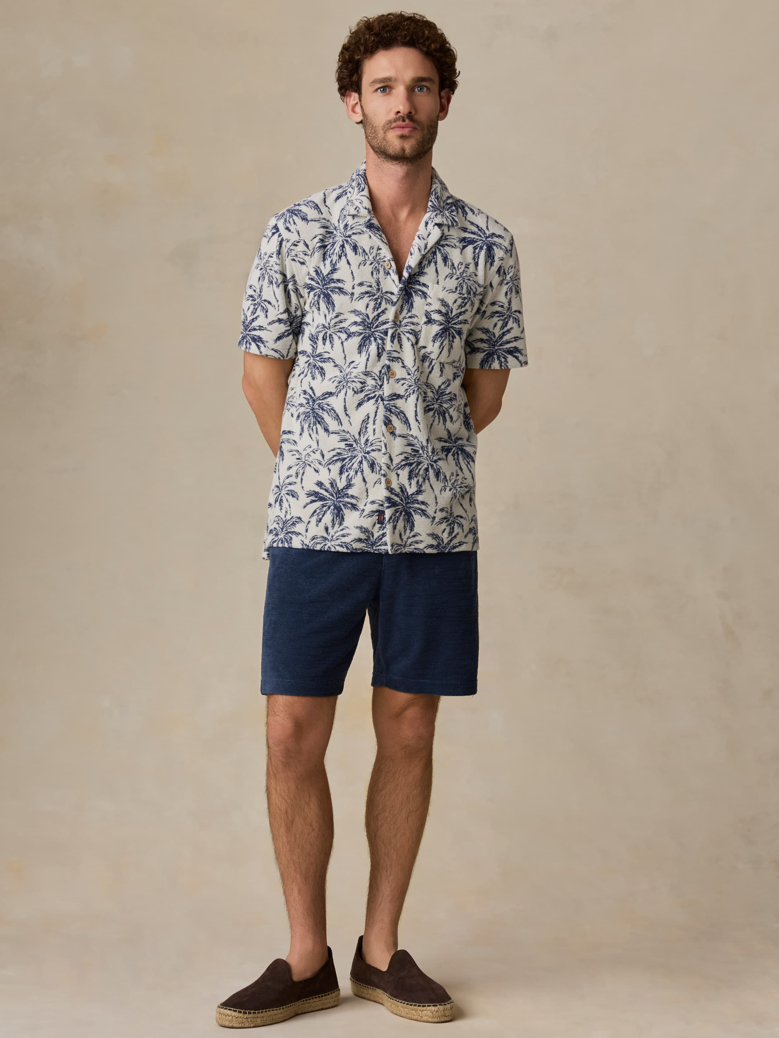 Man wearing a patterned shirt and navy shorts against a beige background