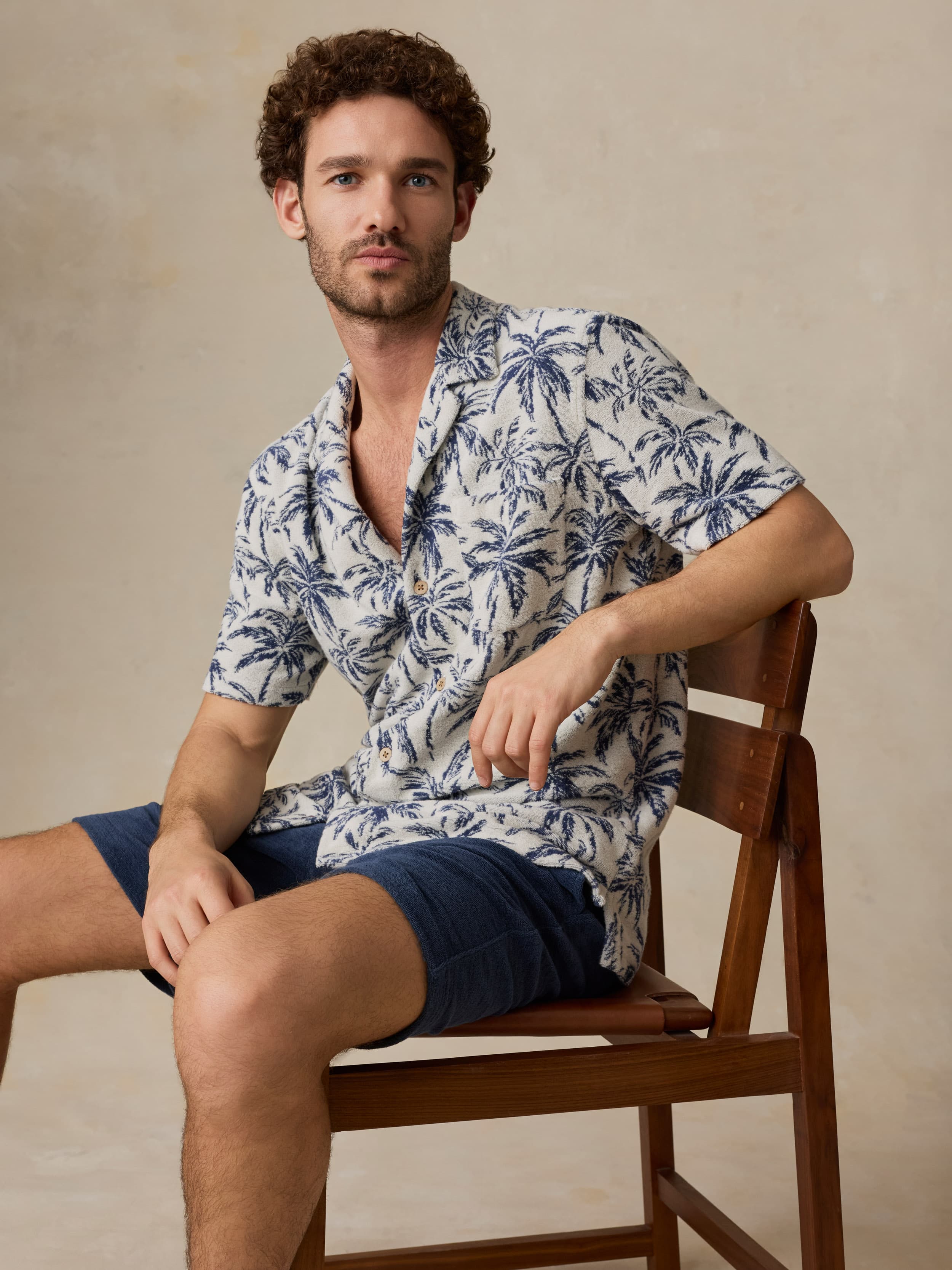 Man wearing a tropical patterned shirt and shorts sitting on a wooden chair against a beige background