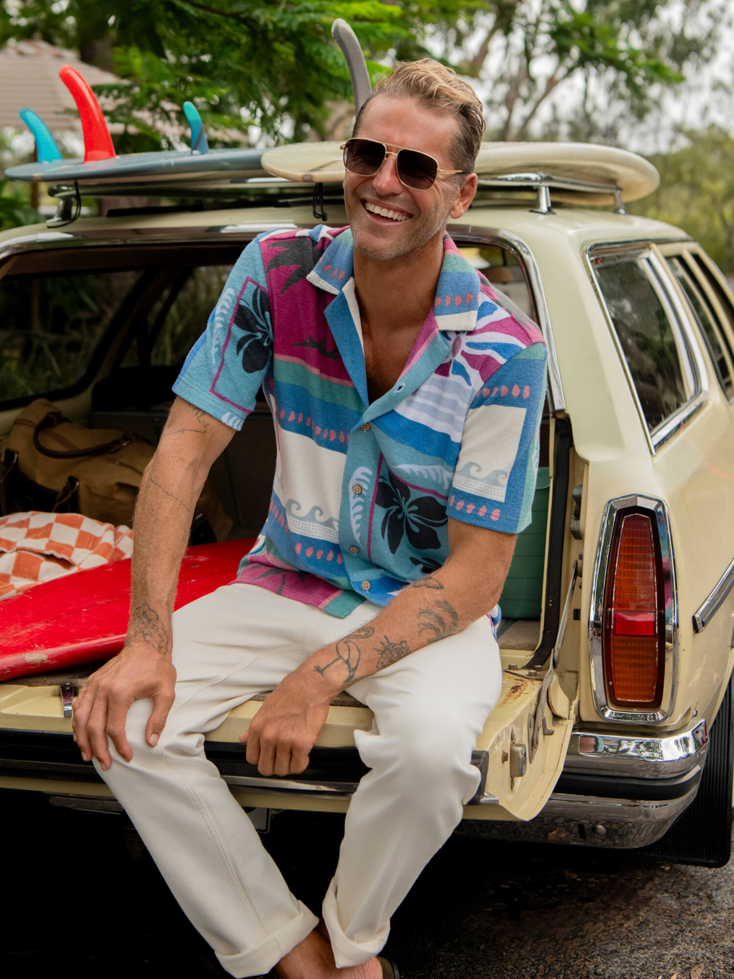 Man wearing a printed short sleeve shirt and white pants sitting in the back of a vintage car