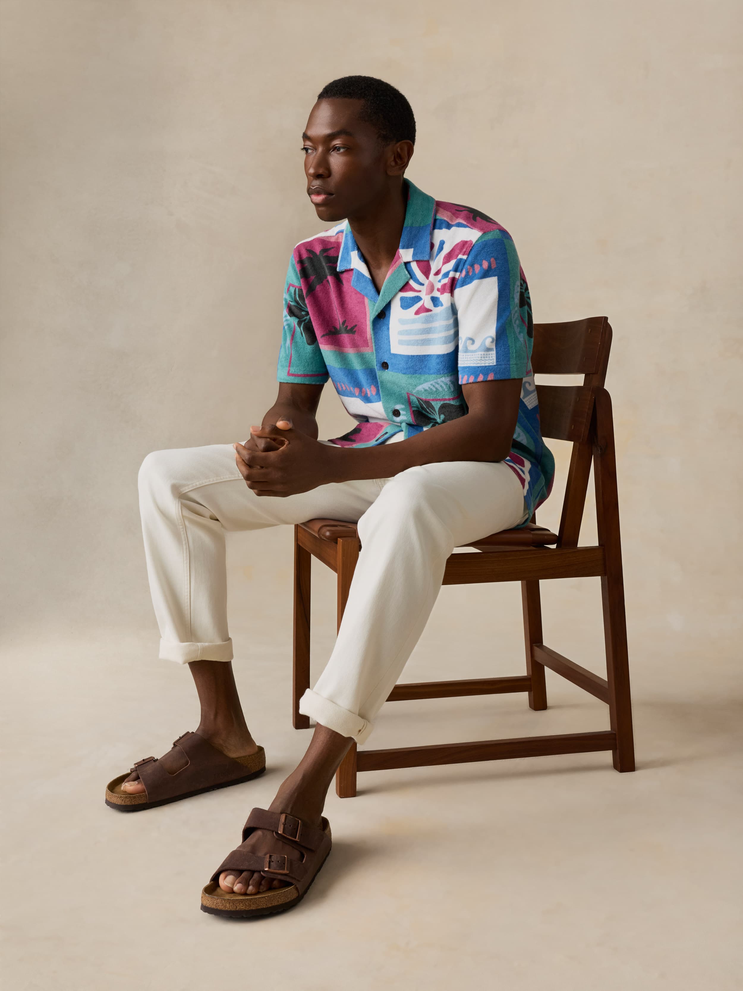 Man sitting on a wooden chair wearing a colorful shirt and white pants.