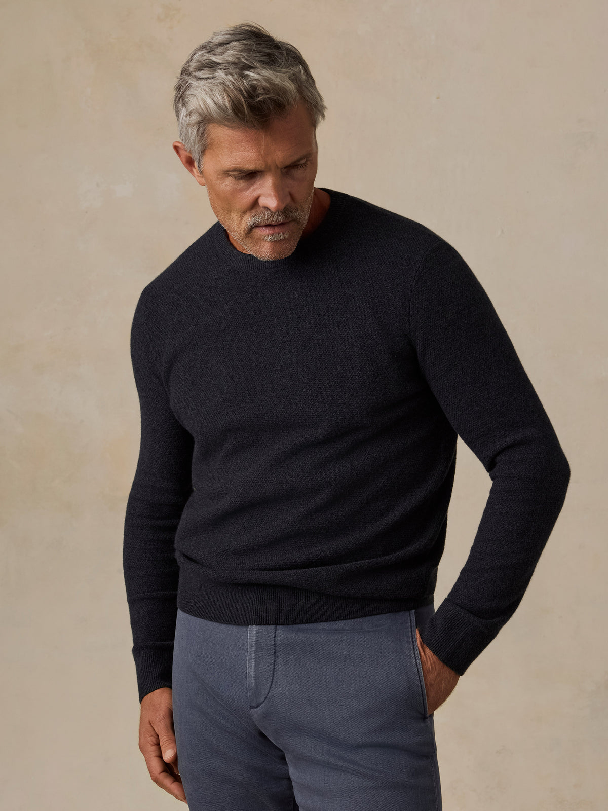 A man wearing a dark navy blue knit crewneck sweater and slate blue trousers, image 1 of 7