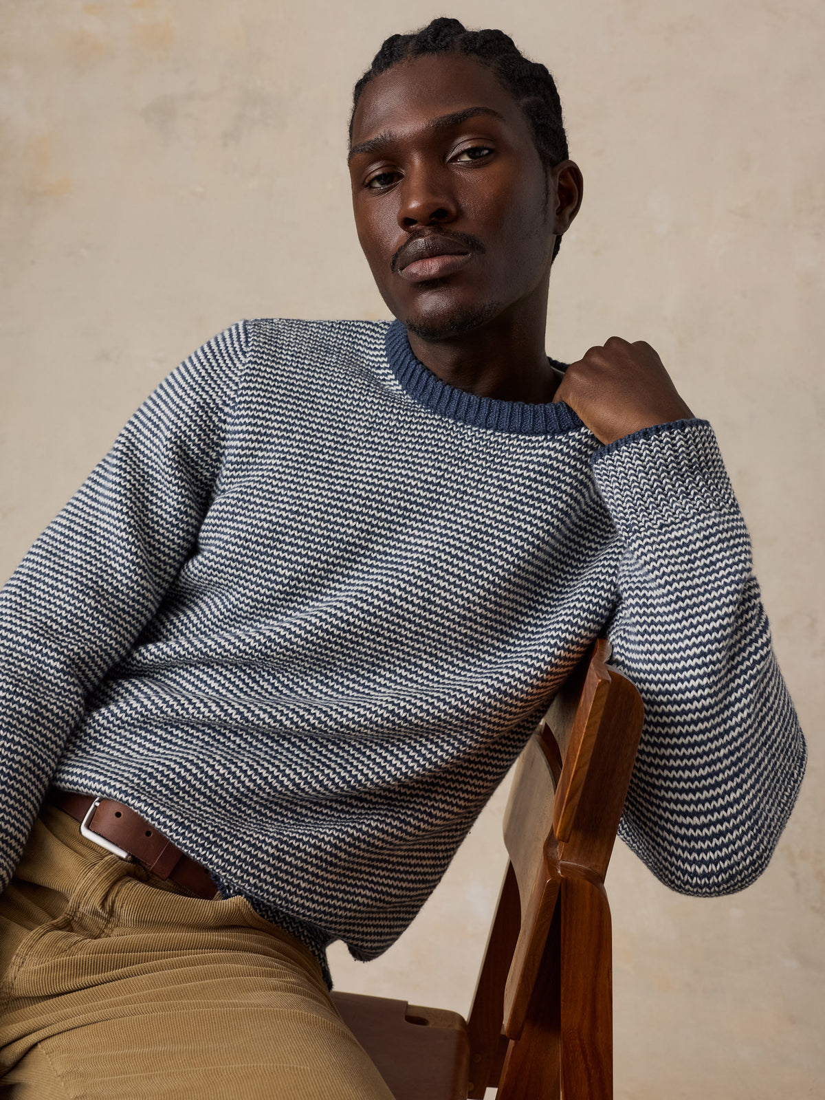 A man sitting on a wooden chair wearing a blue striped sweater and tan corduroy pants, image 4 of 5