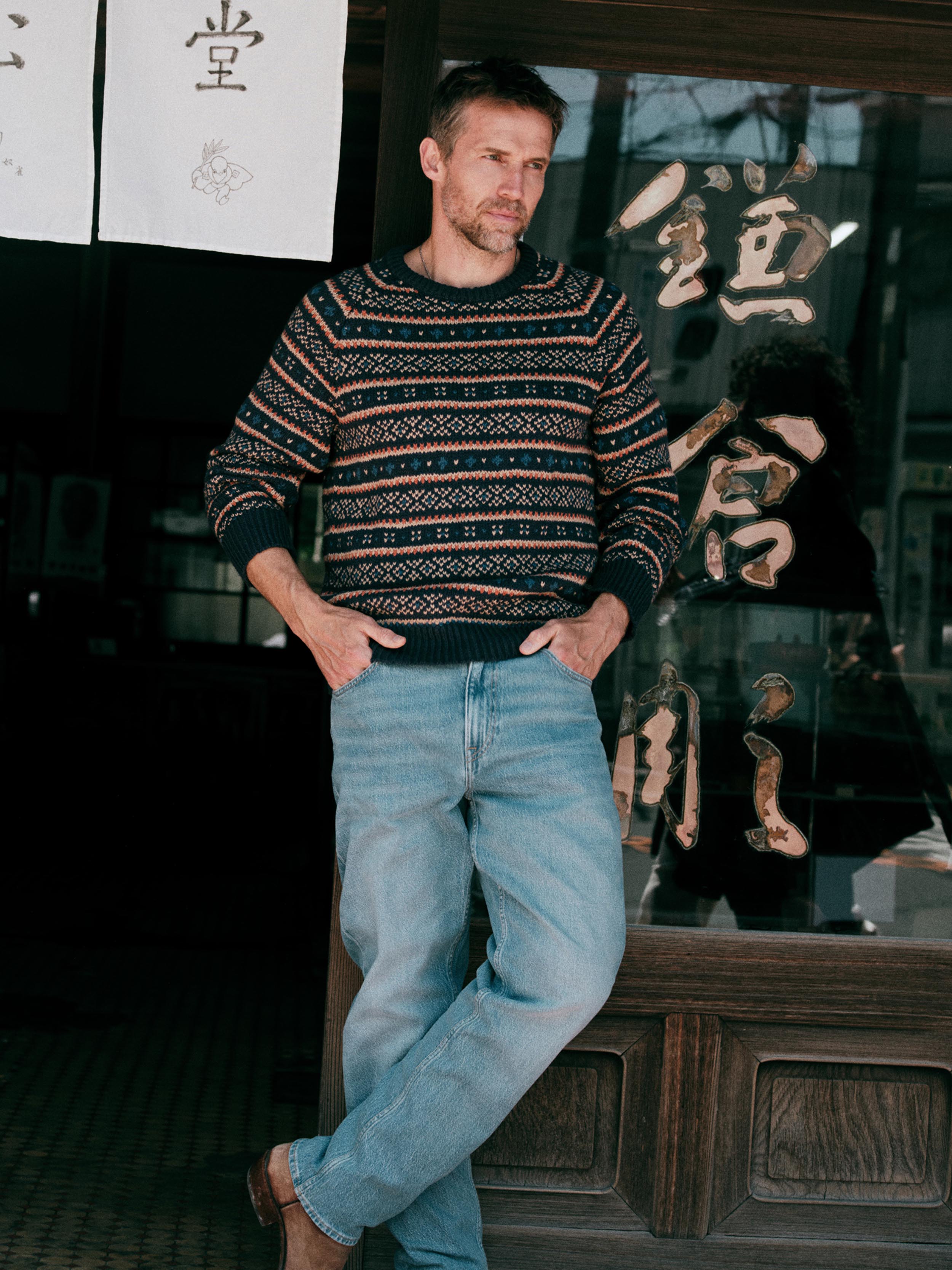 Man wearing a knit fairisle sweater and blue jeans standing outside of a Japanese store
