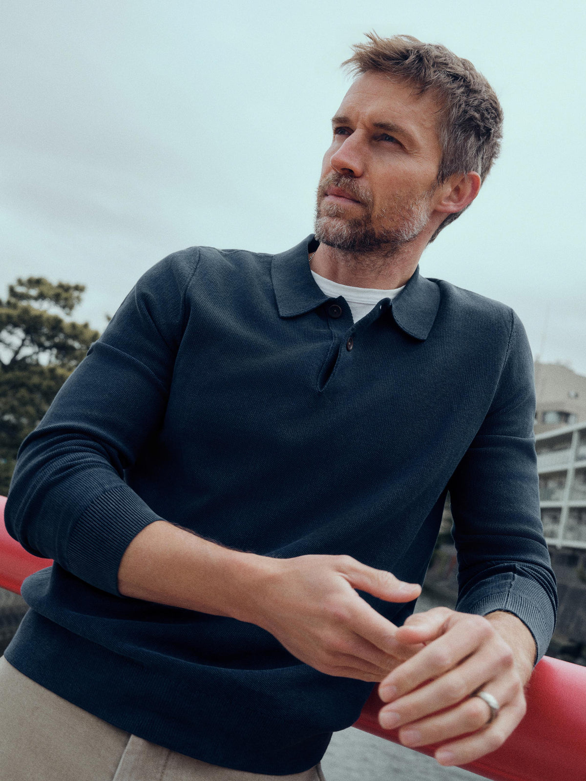A man wearing a dark navy long-sleeve polo sweater over a white undershirt, leaning against a red railing outdoors under an overcast sky., image 1 of 6