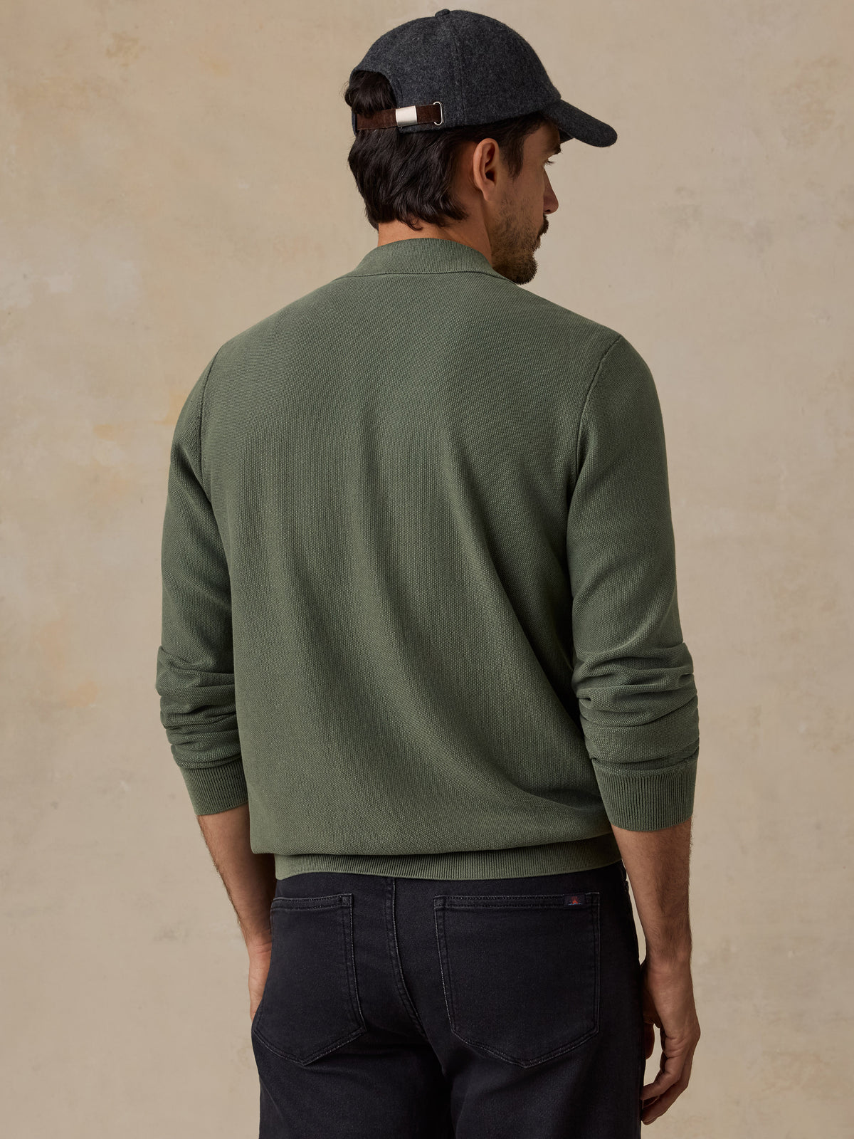 A man shown from the back wearing an olive green knit sweater, dark navy pants, and a dark gray baseball cap, set against a textured beige background., image 3 of 7