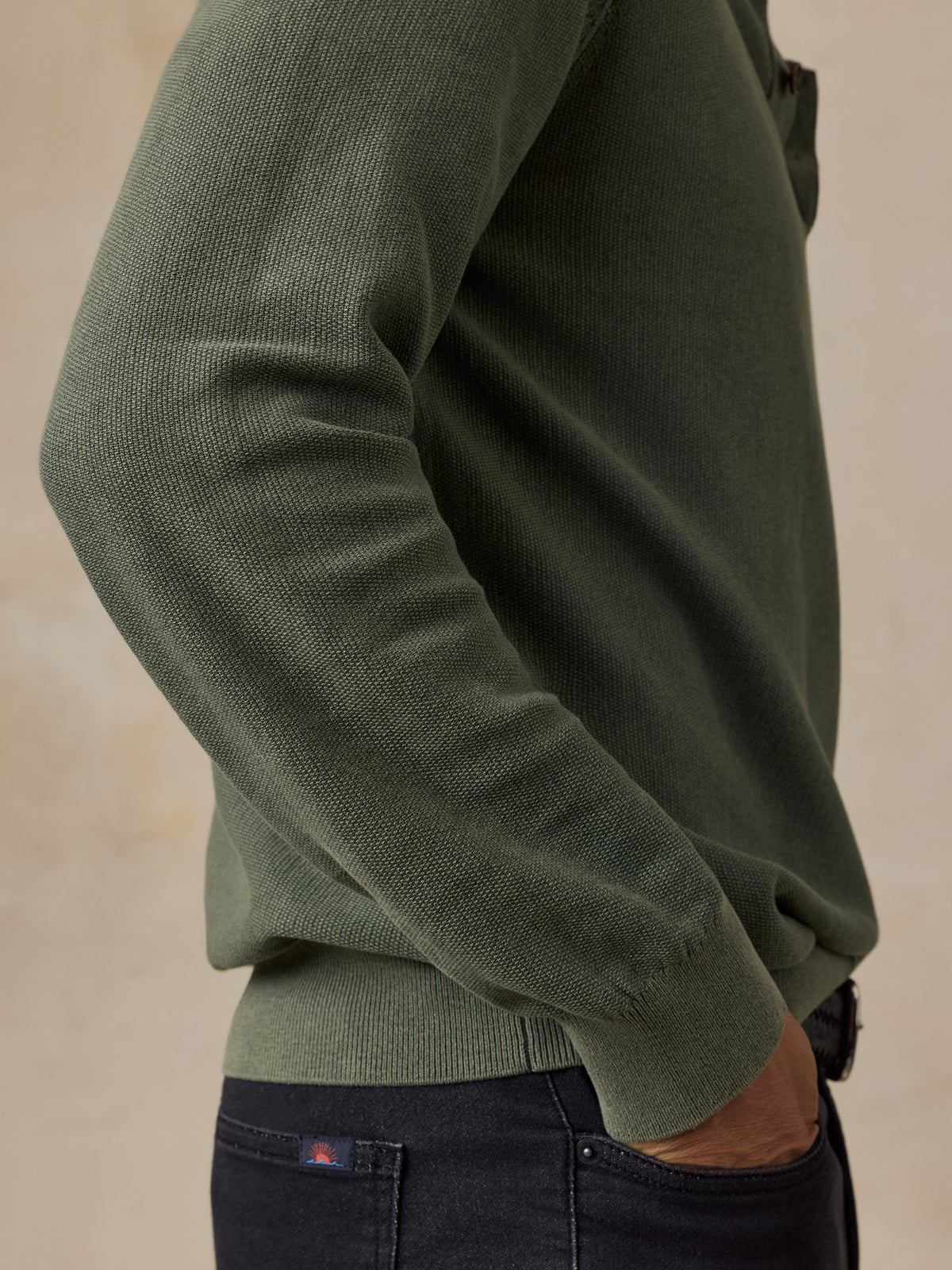 A close-up side view of a man wearing an olive green ribbed knit sweater and dark navy jeans against a warm beige background., image 6 of 7