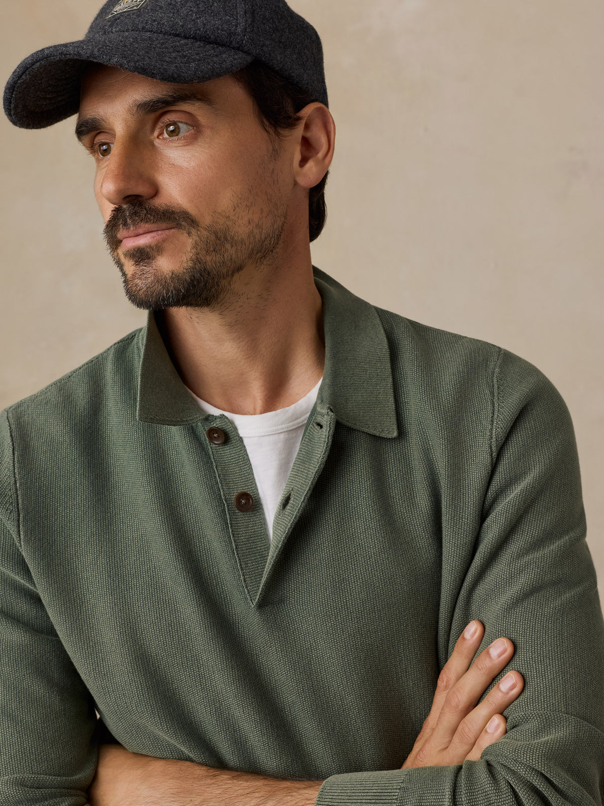 A man wearing an olive green knit polo henley sweater over a white t-shirt, paired with a dark gray baseball hat, set against a warm beige textured background., image 2 of 7